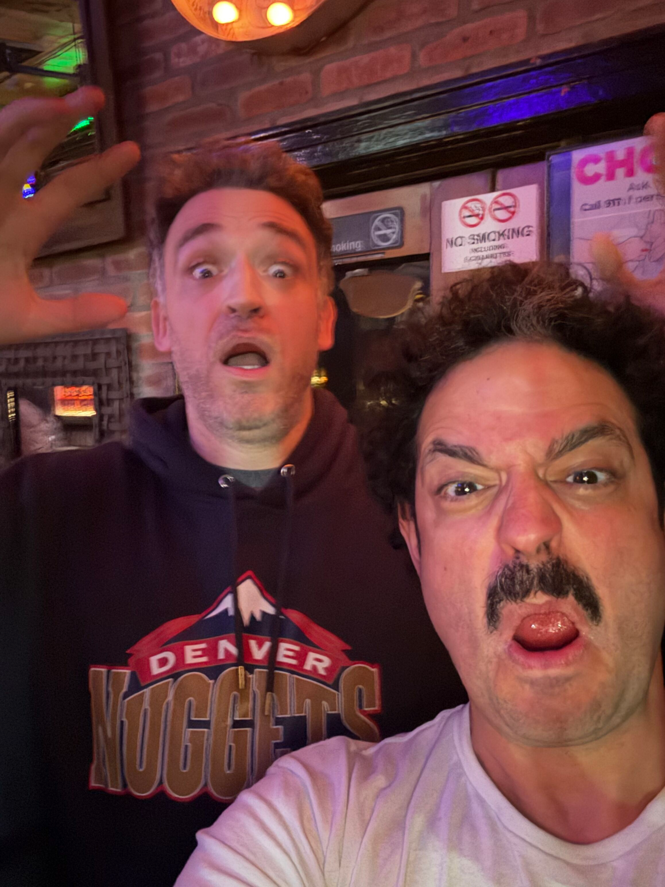 Two men taking a selfie inside a bar or restaurant. One man in a white t-shirt is visible in the foreground with a surprised or playful expression. The other man in the background has wide eyes and an open mouth, raising his hand. The background incl