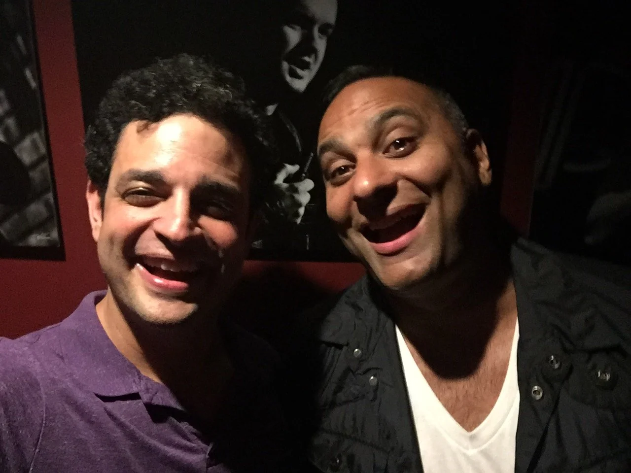 Two men smiling and taking a selfie in a dimly lit room with a black and white photo of a singer in the background.