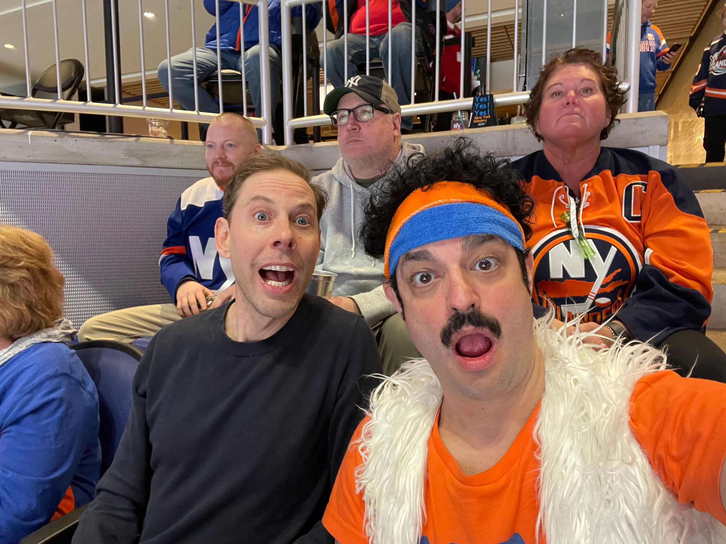 Group of people, some wearing New York Islanders and New York Islanders jerseys, sitting in stadium seats at an indoor arena, with some standing in the background.
