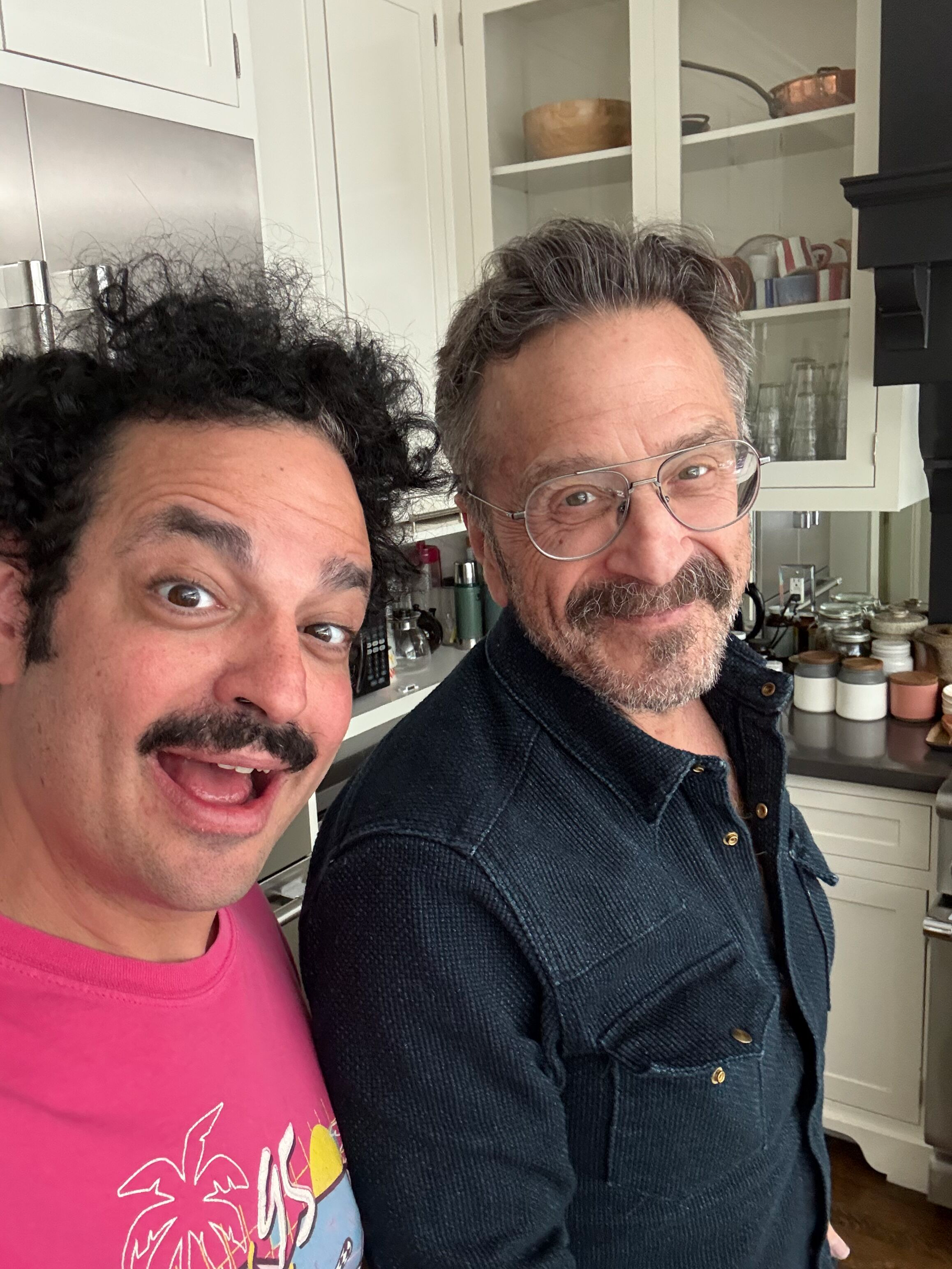 A close-up of two people's faces, a woman on the left wearing a pink shirt with a colorful graphic, and a man on the right wearing glasses and a dark textured jacket, standing in a kitchen with white cabinets and various kitchen items in the backgrou