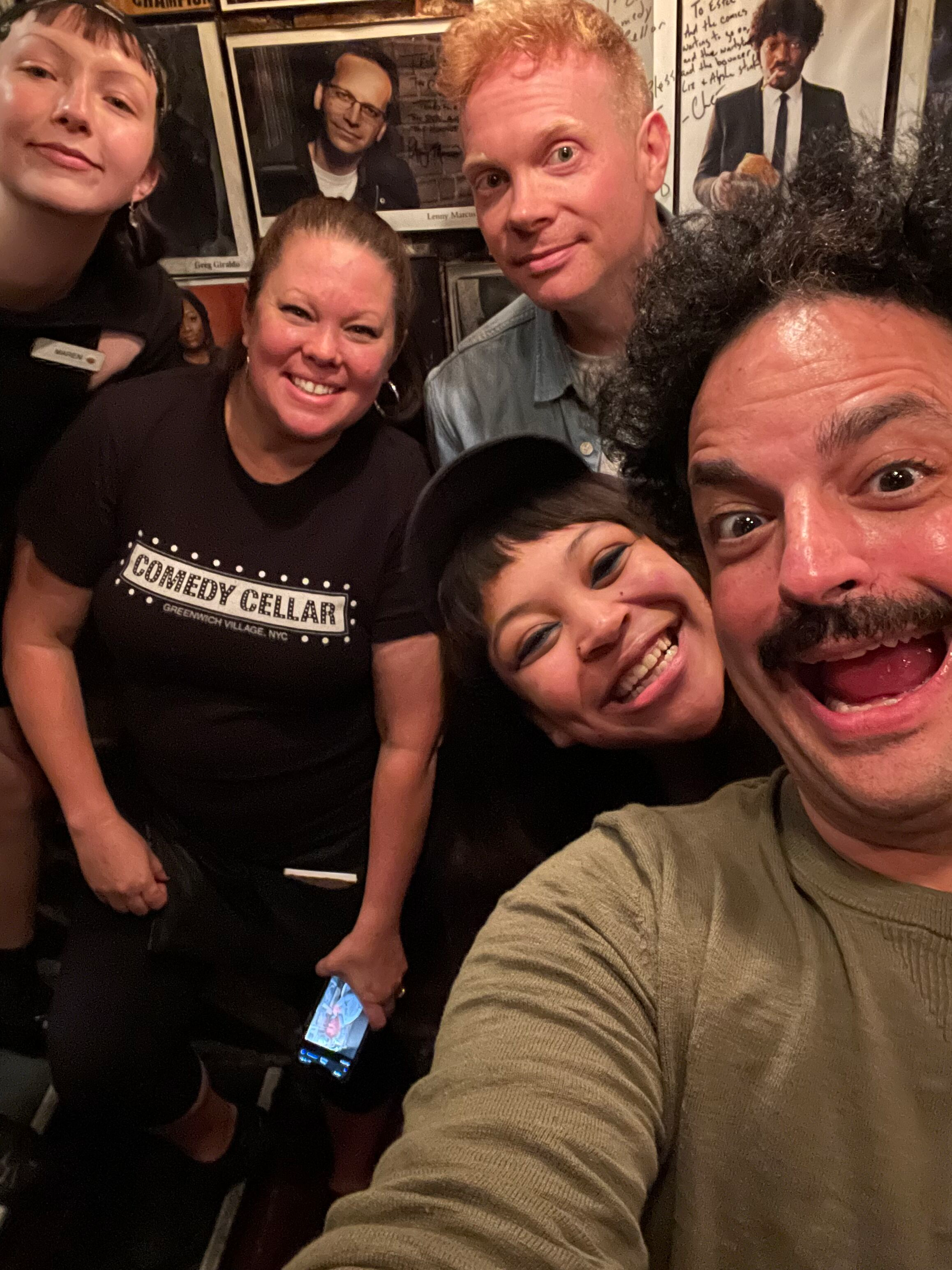 A group of six people taking a selfie in a bar or restaurant, with framed photos of famous musicians on the wall behind them. They are smiling, and one person is holding a smartphone.