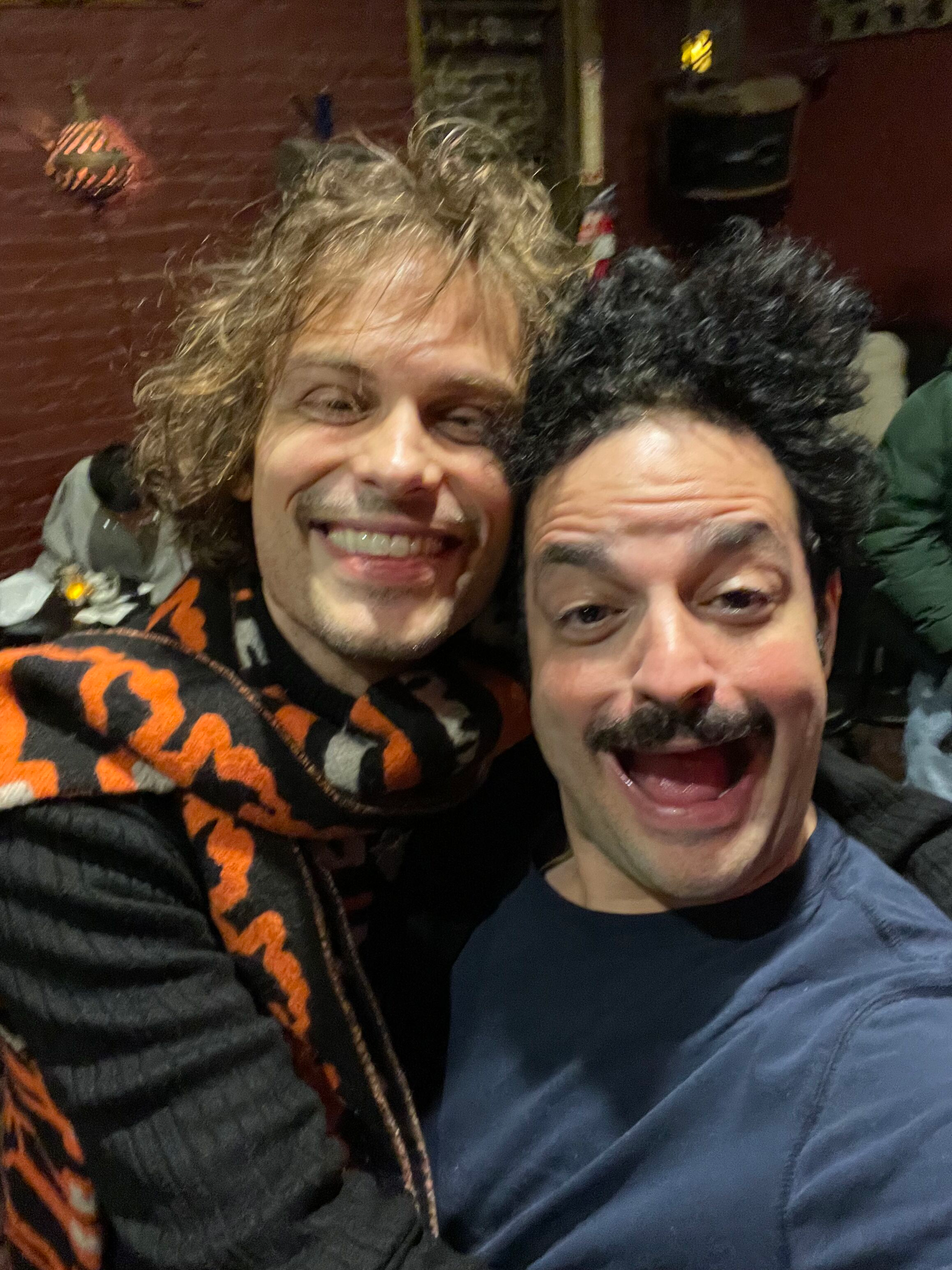 Two smiling men taking a selfie together, one with curly blonde hair and the other with black curly hair, indoors with a brick wall background.