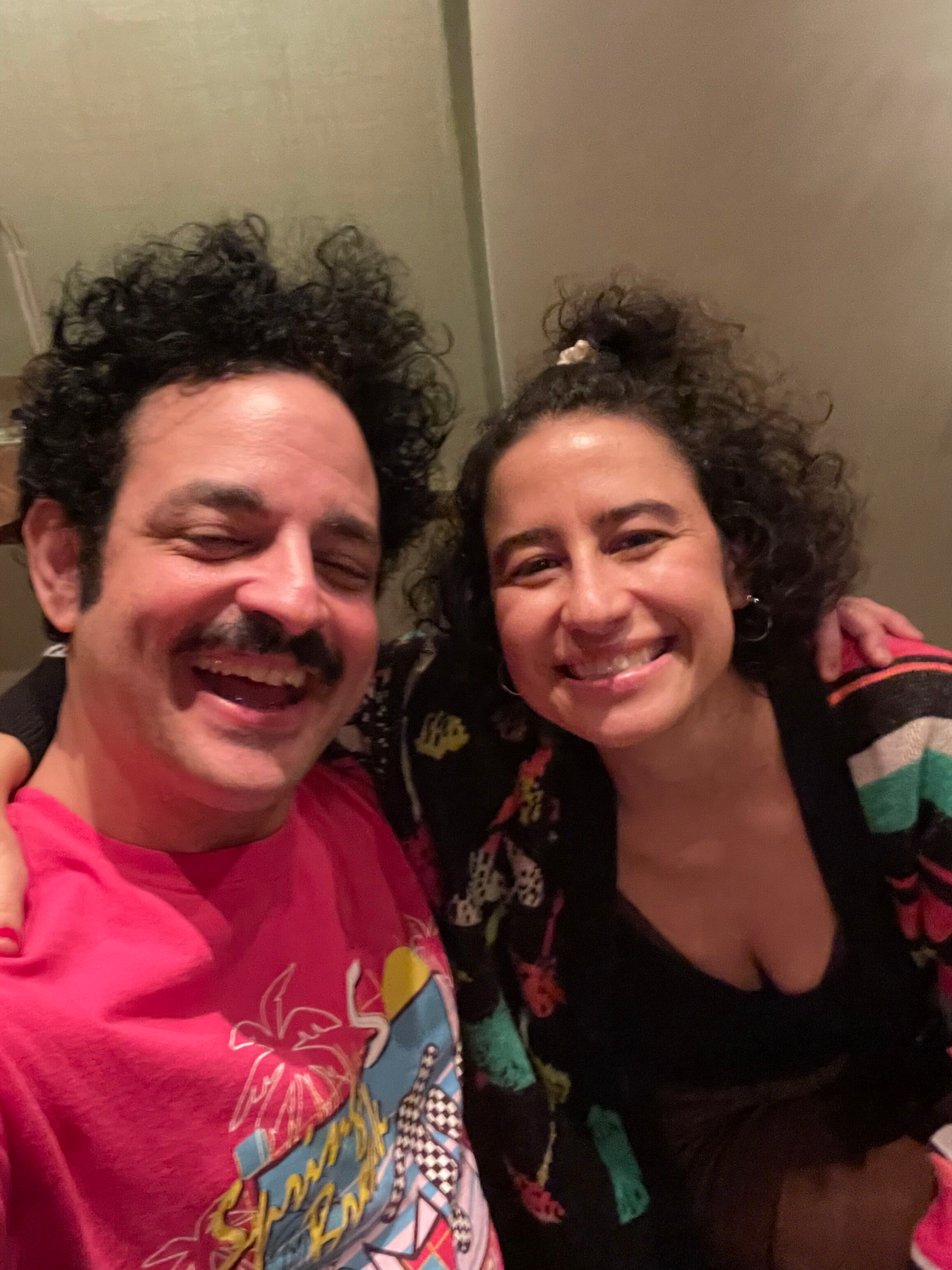 Two people smiling and taking a selfie indoors, with curly hair and casual clothing.