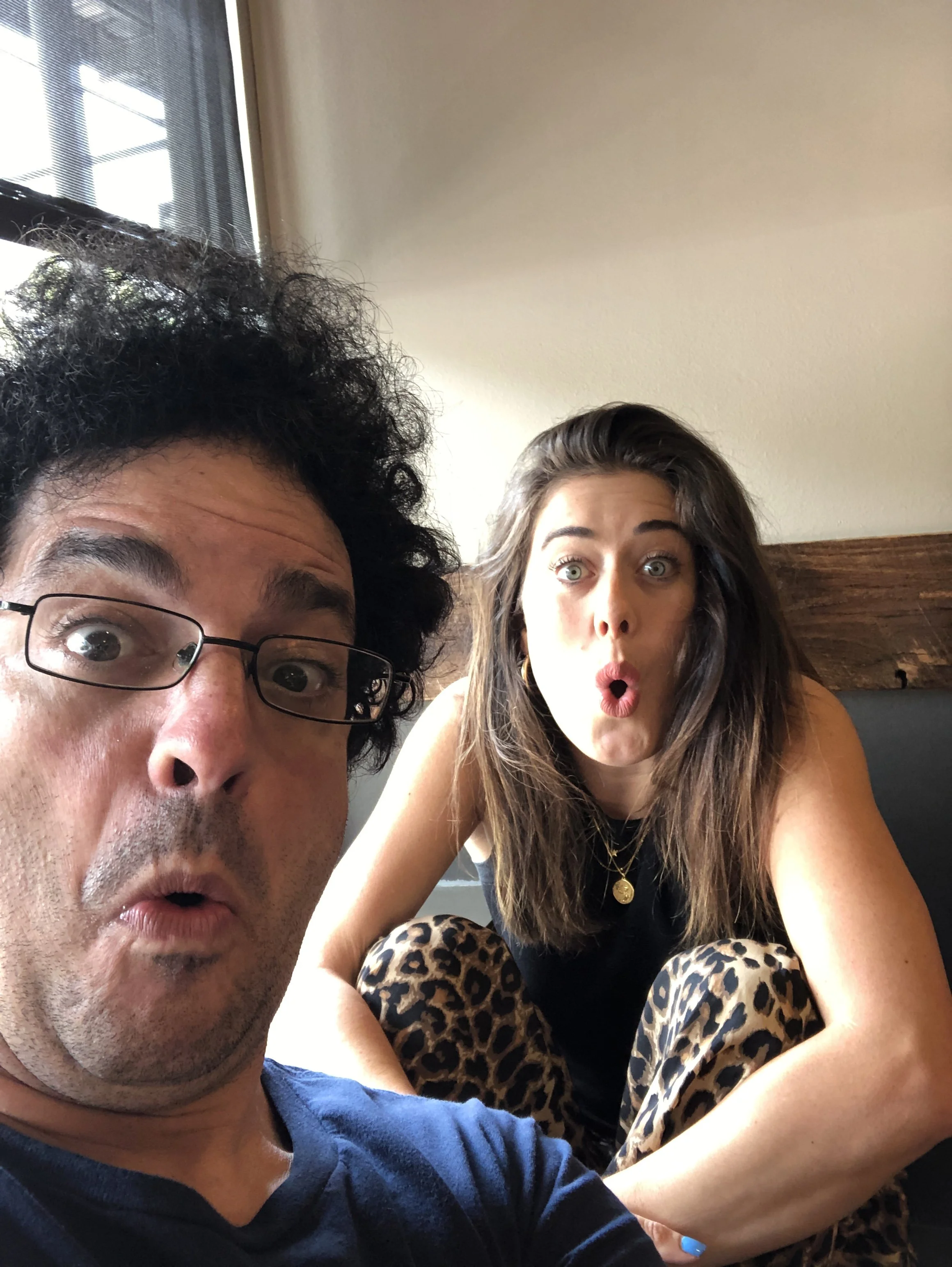 Two people taking a selfie, a man with glasses and curly hair on the left and a woman with wavy hair and blue eyes on the right, both making surprised facial expressions, inside a room with a window and wooden wall.