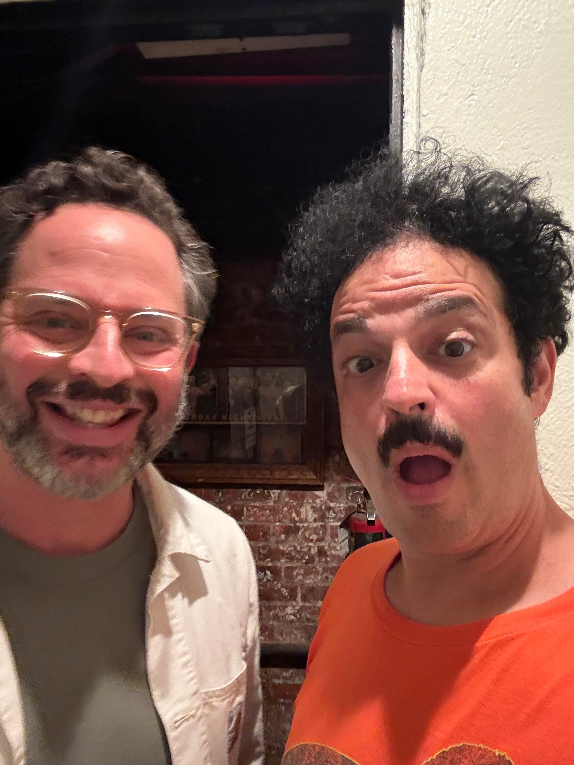 Two smiling men taking a selfie together, one wearing glasses and a beige shirt, the other with dark, curly hair and an orange shirt, standing inside a room with a brick wall and a painting in the background.