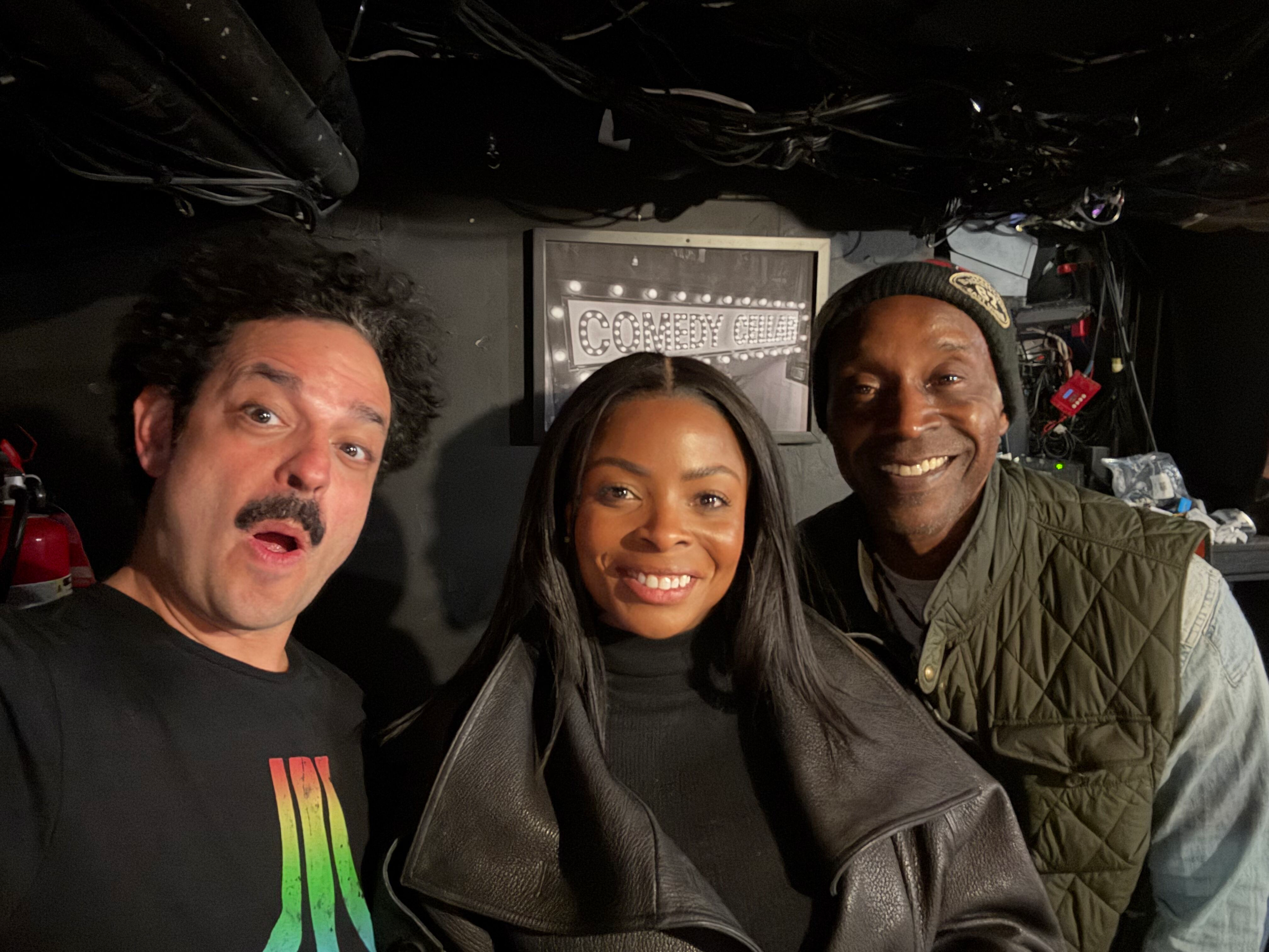 Three smiling people taking a selfie together in a comedy club, with a stage and sign that says 'Comedy