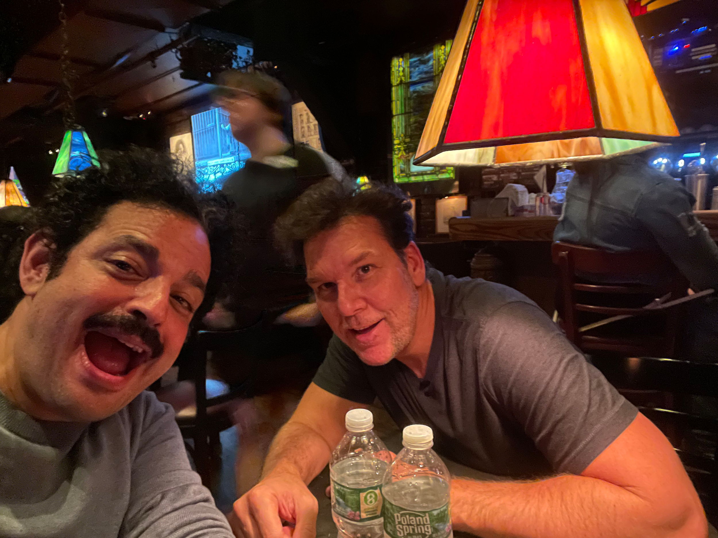 Two men taking a selfie in a dimly lit restaurant or bar, with colorful stained glass lamps on the ceiling and bottles of Poland Spring water on the table.