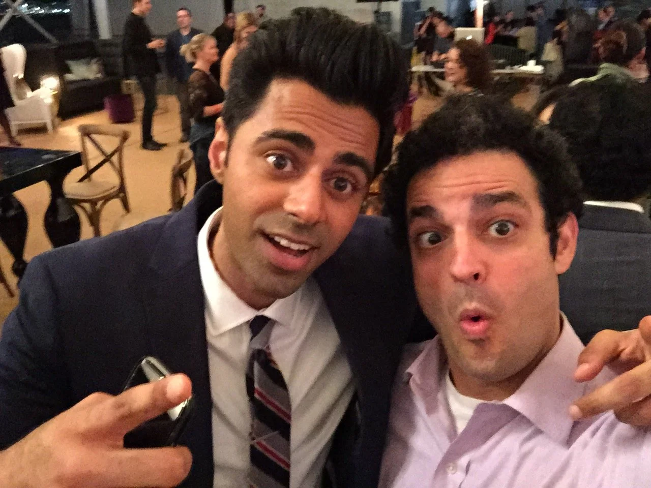 Two men taking a selfie at a social event. The man on the left is wearing a dark suit and tie, and the man on the right is wearing a pink dress shirt. They are both making surprised or excited facial expressions. The background shows other people soc