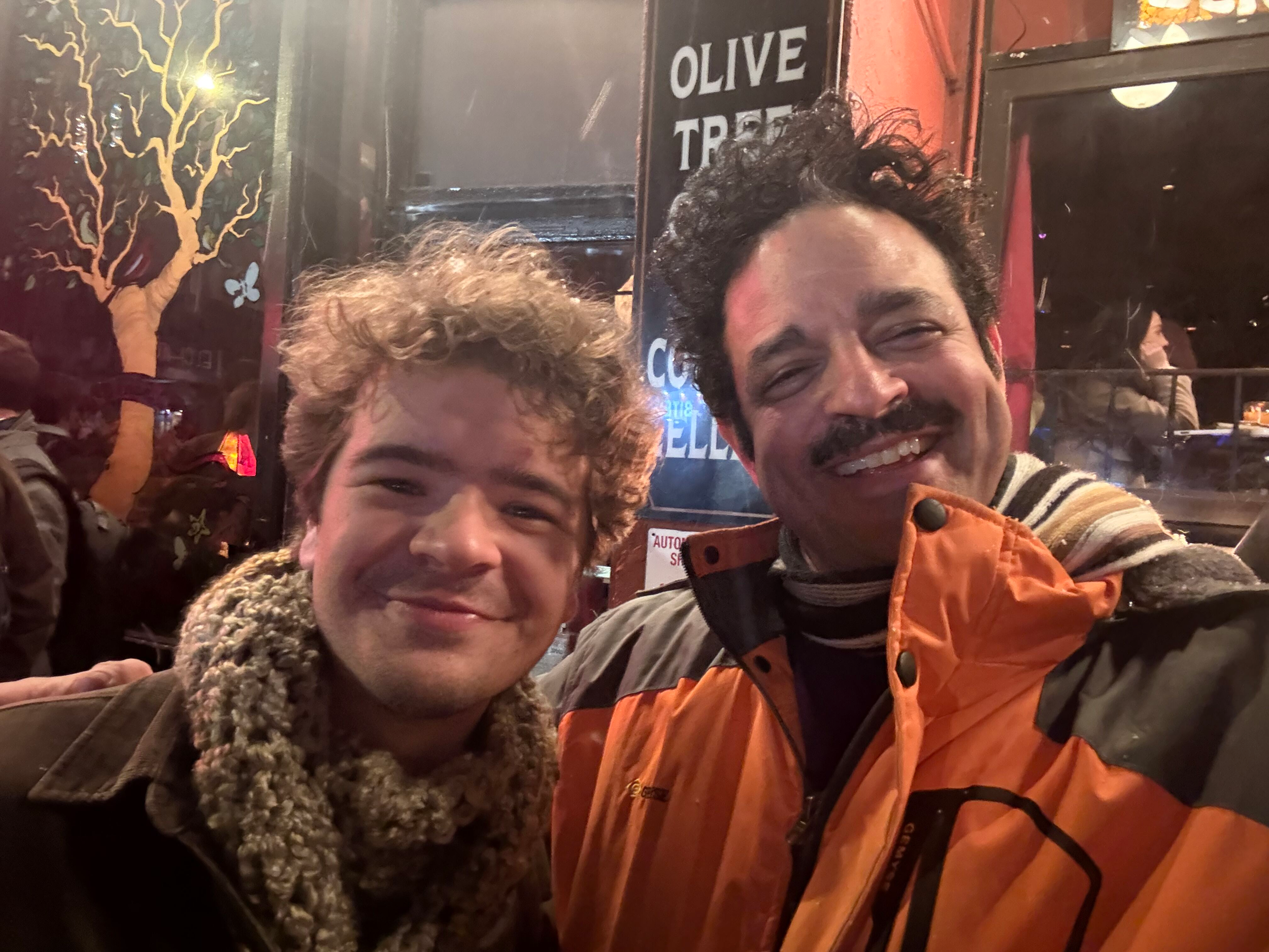 Two smiling men taking a selfie at night outside a restaurant with a sign that reads 'Olive Tree'. There are other people in the background and a tree painted on the wall.