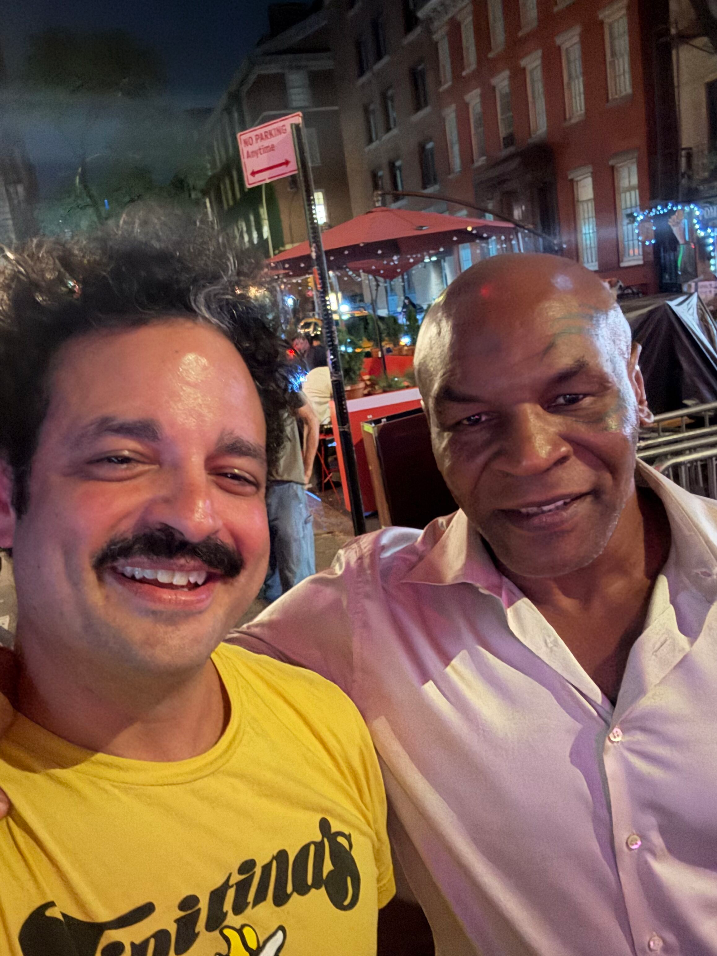Two men smiling and taking a selfie outdoors at night, with city buildings and festive decorations in the background.