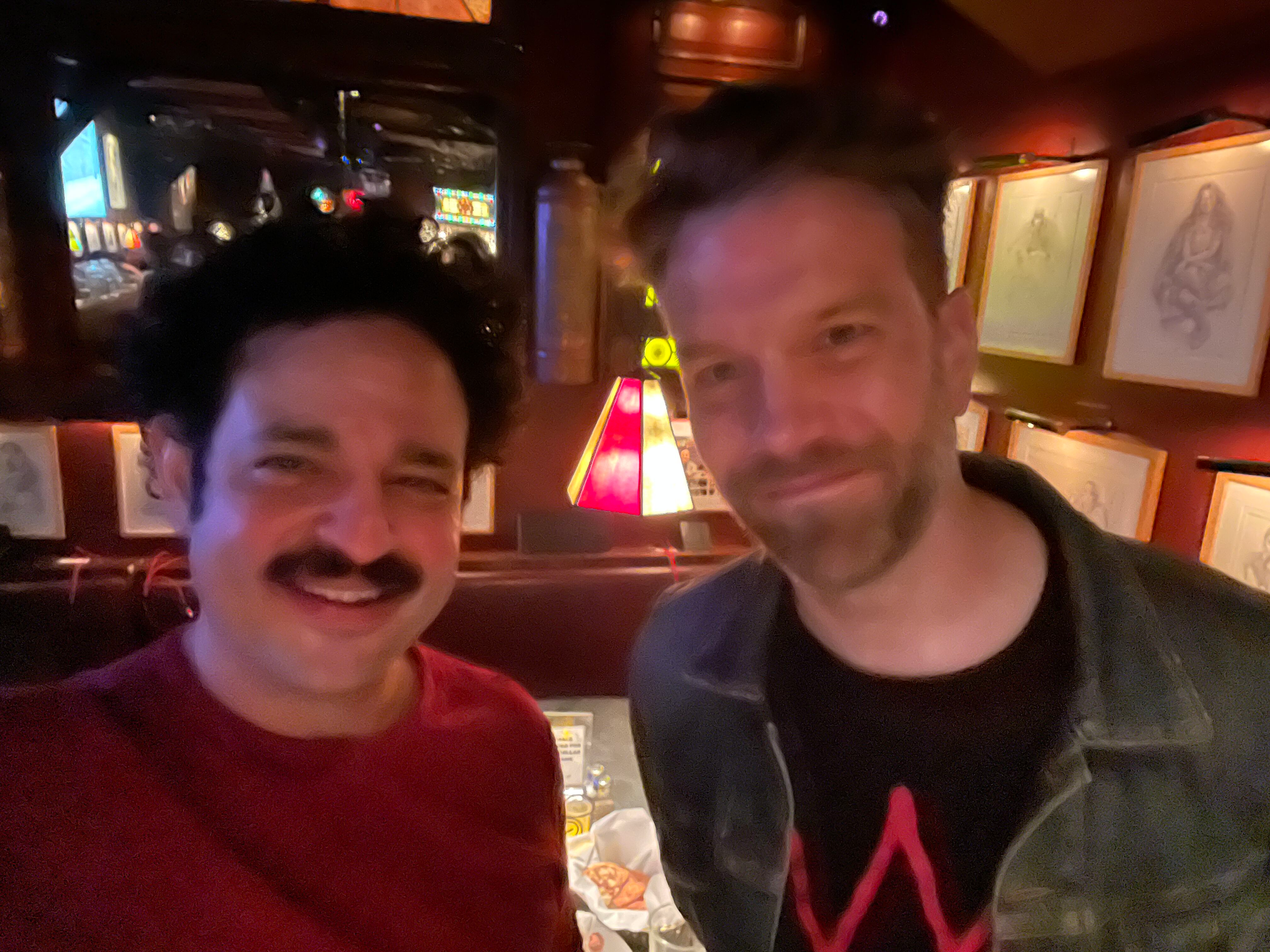 Two men smiling for a photo in a dimly lit restaurant or bar with framed artwork on the wall behind them.