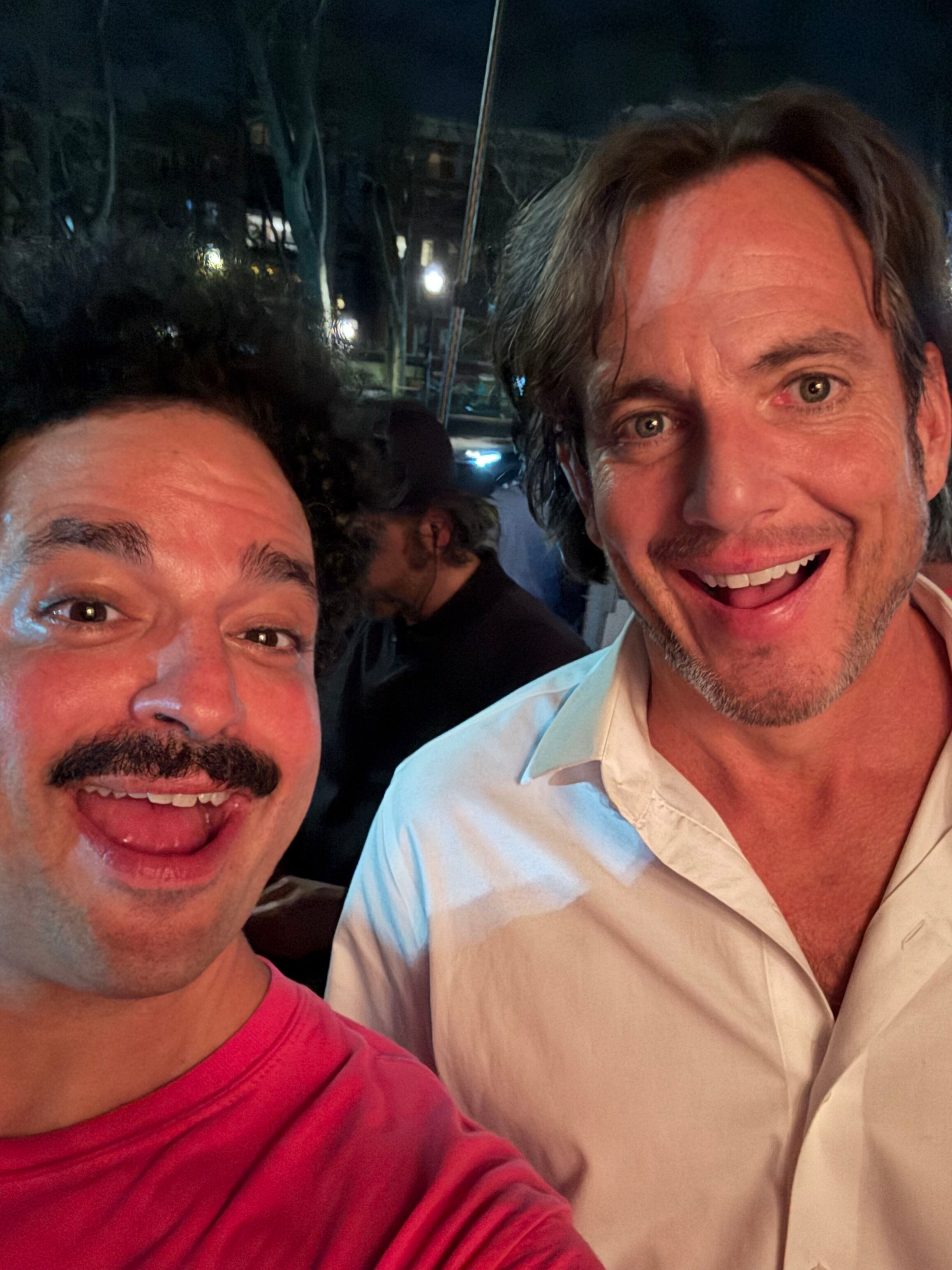 Two men taking a selfie, smiling, with dark hair, one with curly hair and a beard, wearing a red shirt, and the other with straight hair, wearing a white shirt, in a dimly lit environment with some lights and a person blurred in the background.
