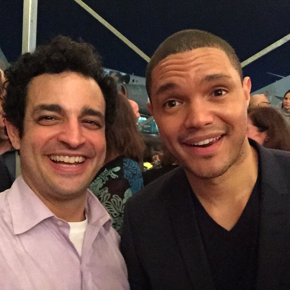Two men smiling for a selfie at an outdoor event with many people in the background.
