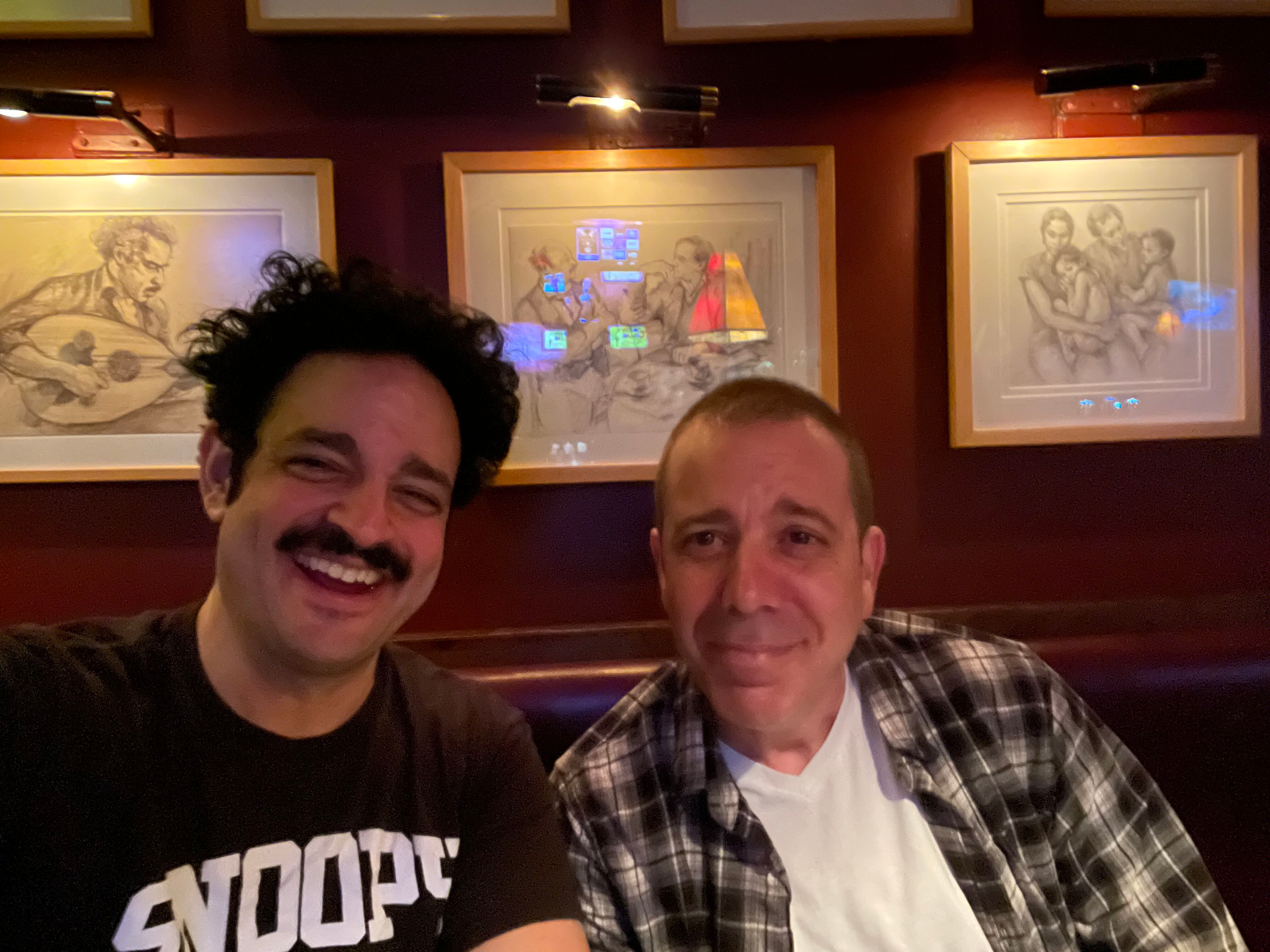 Two men smiling for a photo in a dimly lit room with framed pictures on the wall behind them. The man on the left has curly dark hair, a mustache, and is wearing a black T-shirt. The man on the right has short hair, is clean-shaven, and is wearing a 