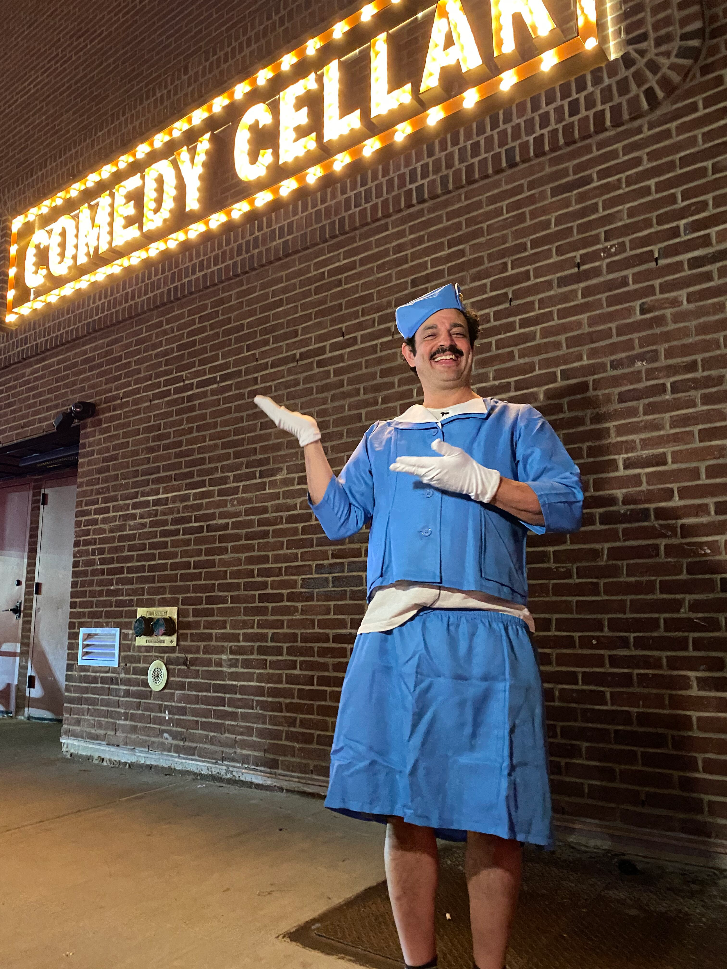 A man dressed in a blue costume resembling a nurse or maid outfit, with a matching cap and white gloves, standing in front of a brick wall with a large, illuminated sign that reads 'Come and get it'. The man is smiling and pointing at the sign.