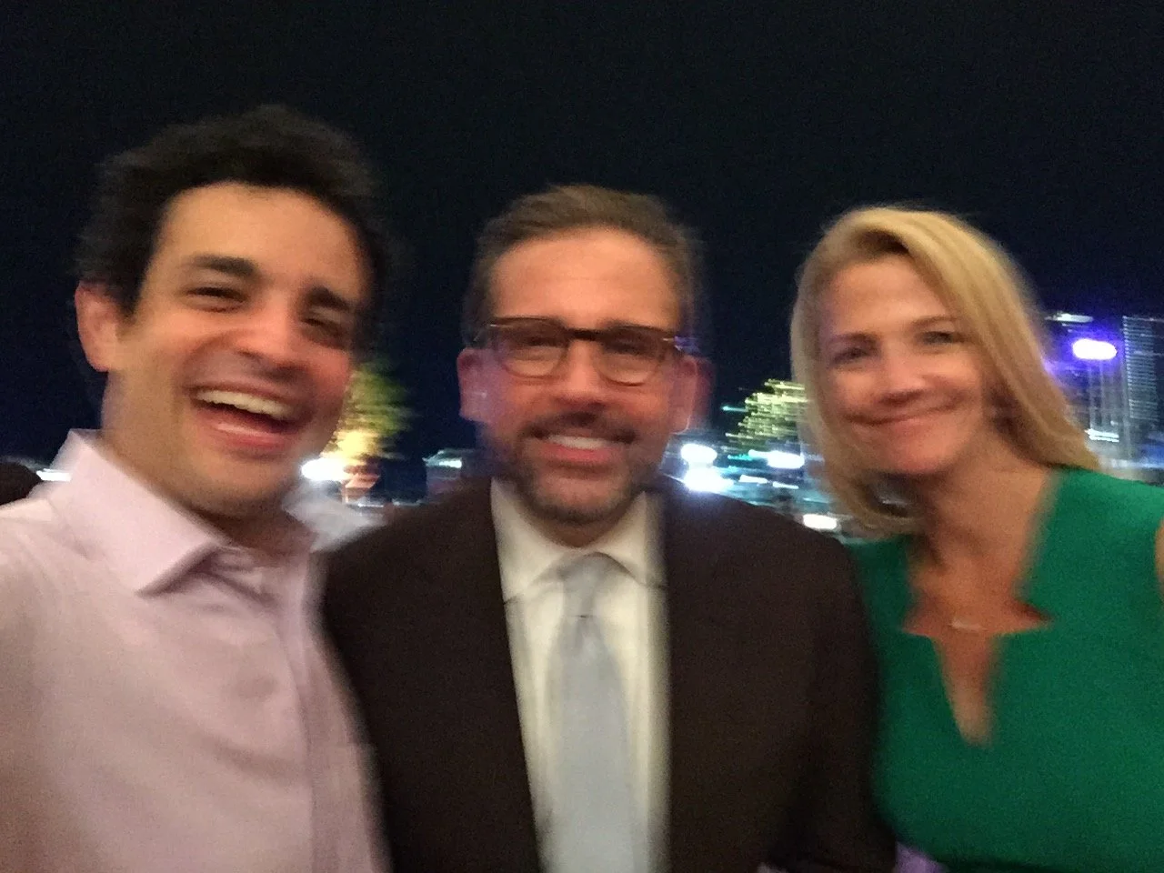 Three people smiling and posing for a photo at night with city lights in the background.