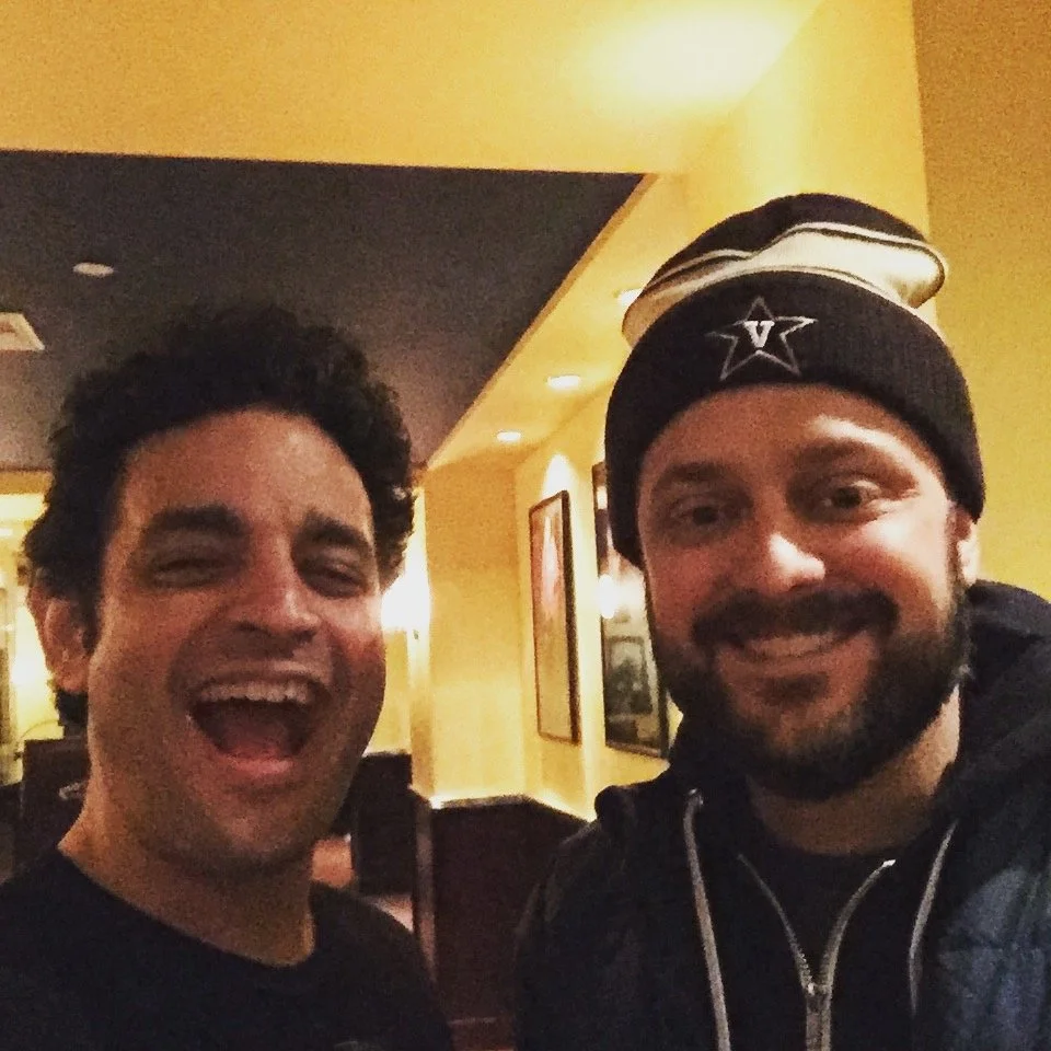 Two smiling men in a restaurant, one with dark curly hair and the other with a beard, wearing a beanie with a star and stripe design.