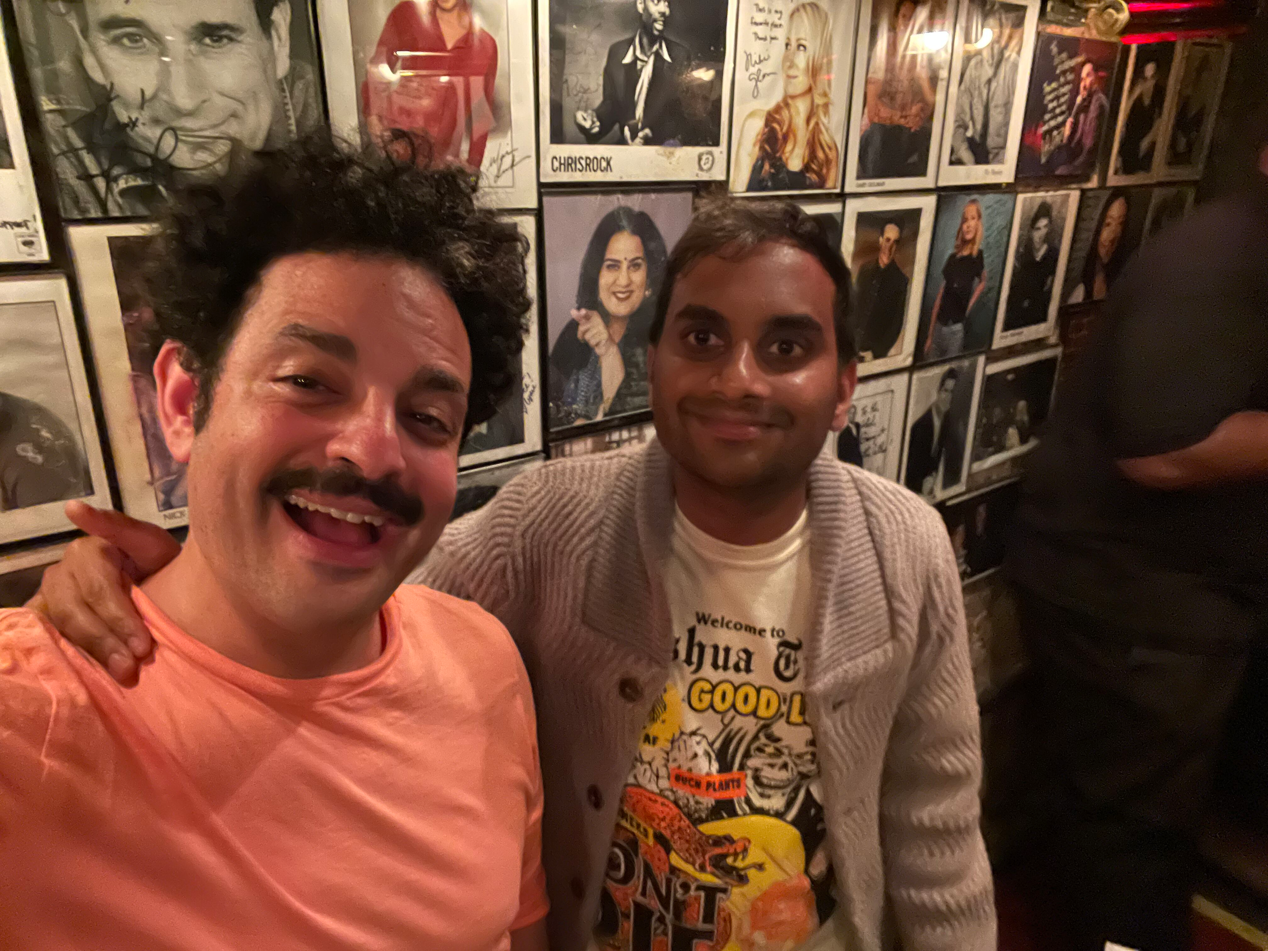 Two men taking a selfie in front of a wall covered with framed photographs of celebrities and signed posters in a dimly lit room.