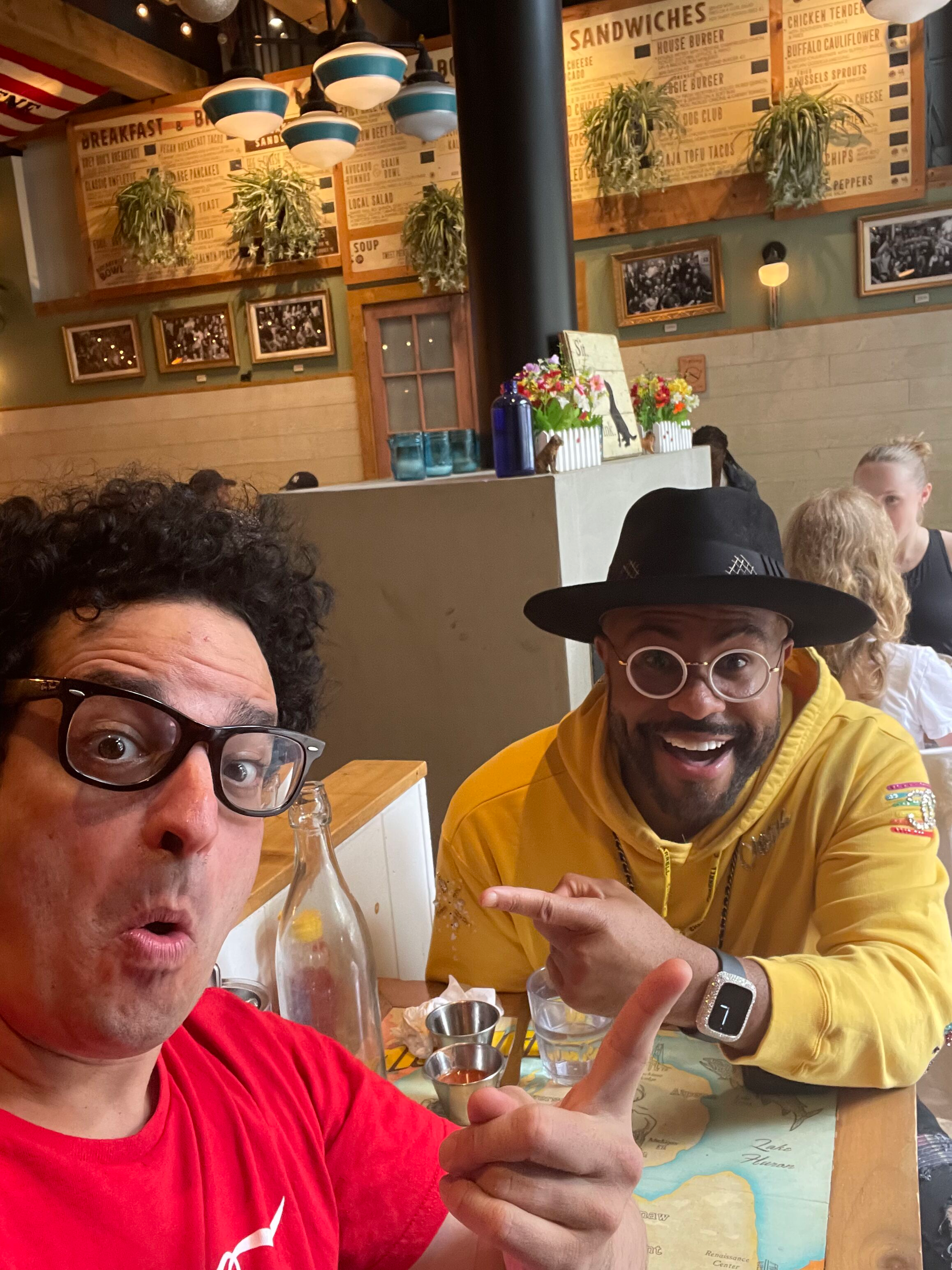 Two men are sitting at a restaurant table, taking a selfie. The man on the left has curly hair, glasses, and is wearing a red shirt. The man on the right has a beard, glasses, and is wearing a yellow hoodie and a black hat. They are smiling and point