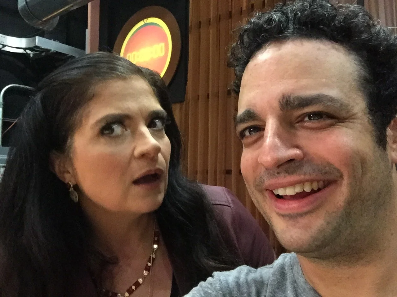 A close-up selfie of a man smiling with a woman in the background looking surprised, indoors with wooden paneling and a sign in the background.