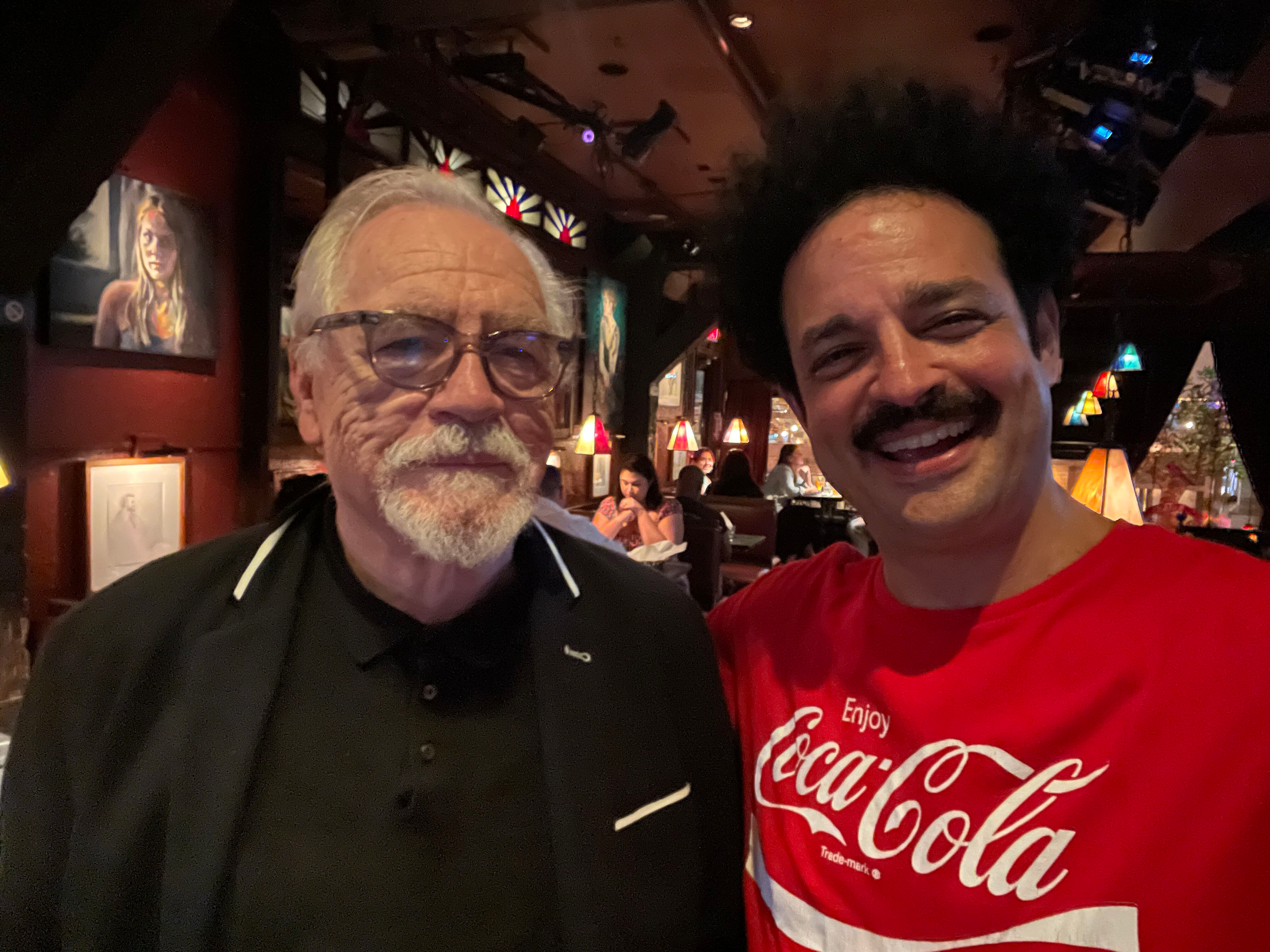 A selfie of two men smiling in a dimly lit restaurant or bar. The man on the left is older with white hair, glasses, and a beard, wearing a black shirt. The man on the right is younger with dark, curly hair, a mustache, and a big smile, wearing a red