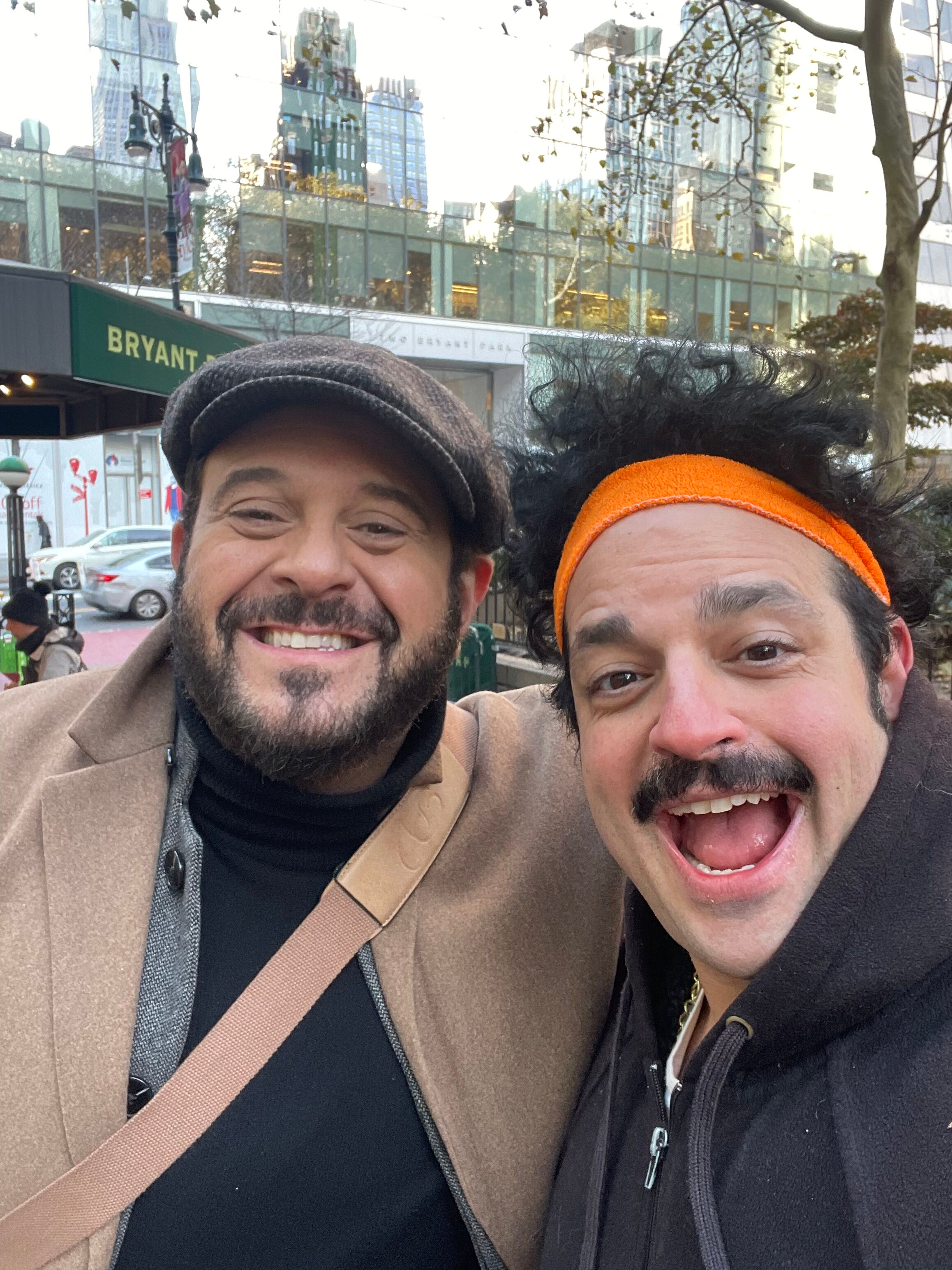 Two smiling men taking a selfie outdoors in a city, with tall buildings and a glass facade in the background.