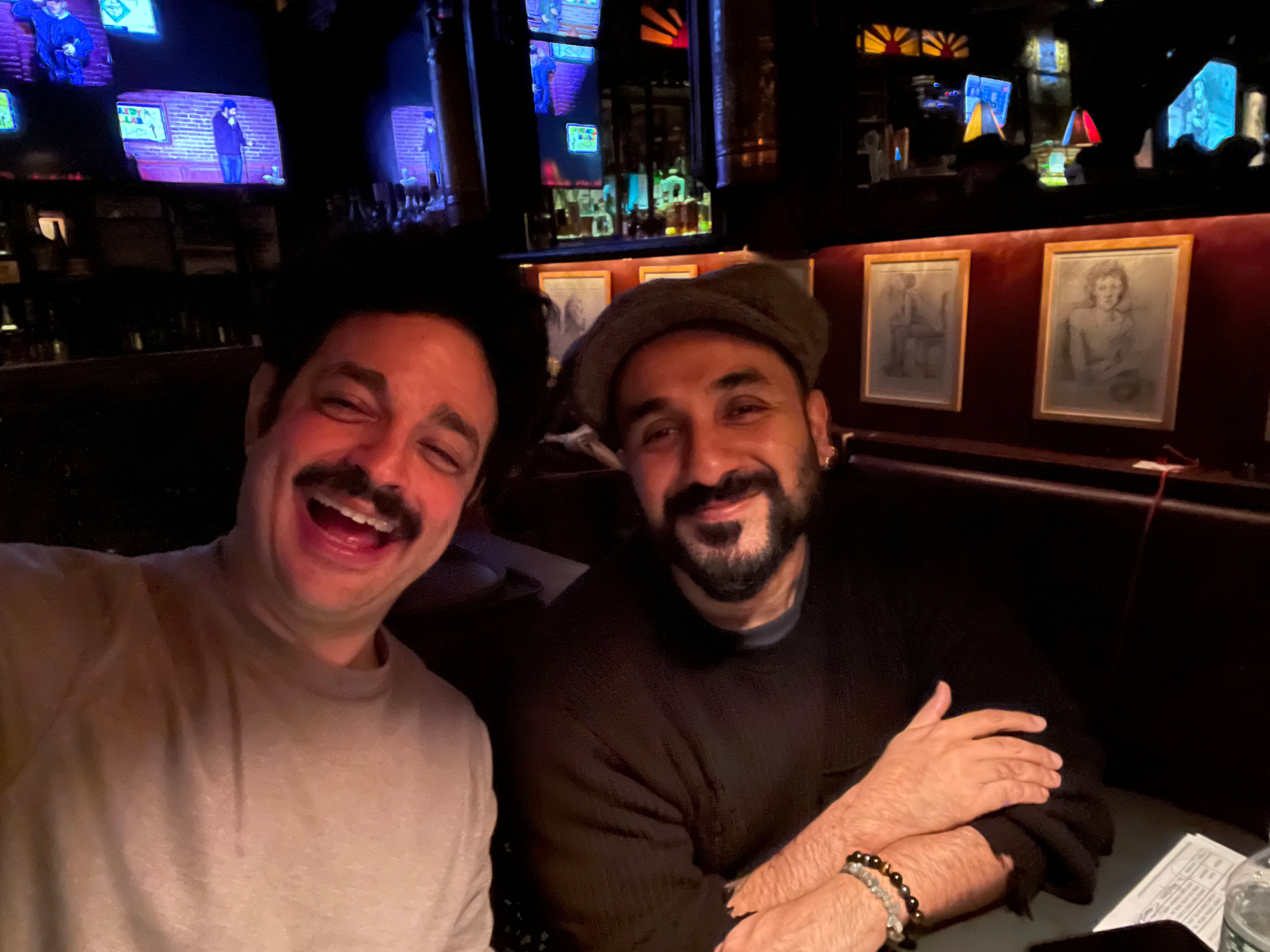 Two smiling men taking a selfie in a dimly lit bar or restaurant, with framed pictures on the red wall behind them and multiple television screens showing a stand-up comedy performance.