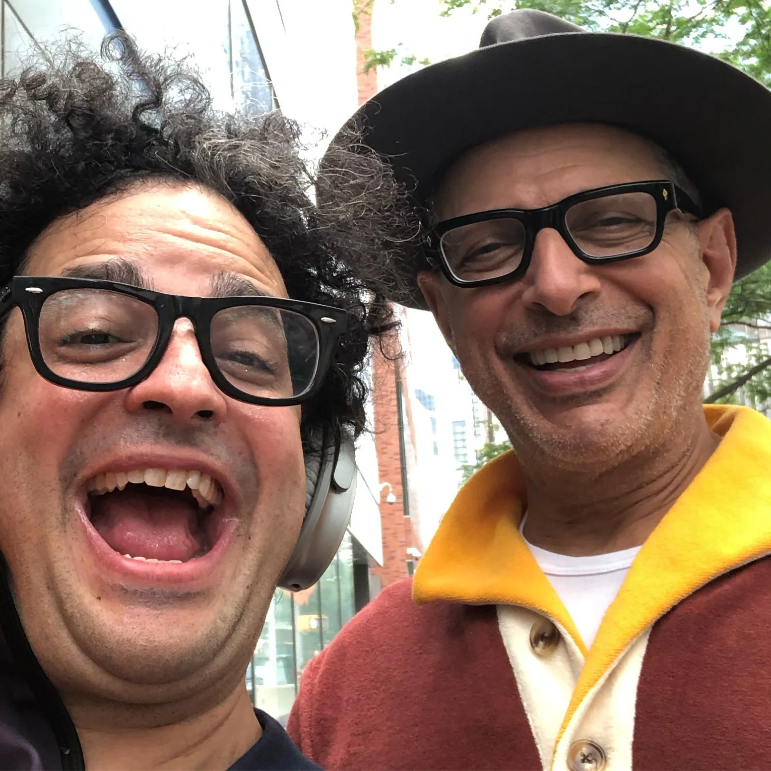 Close-up selfie of two smiling men wearing glasses, one with curly hair and the other with a hat, outdoors with trees and buildings in the background.