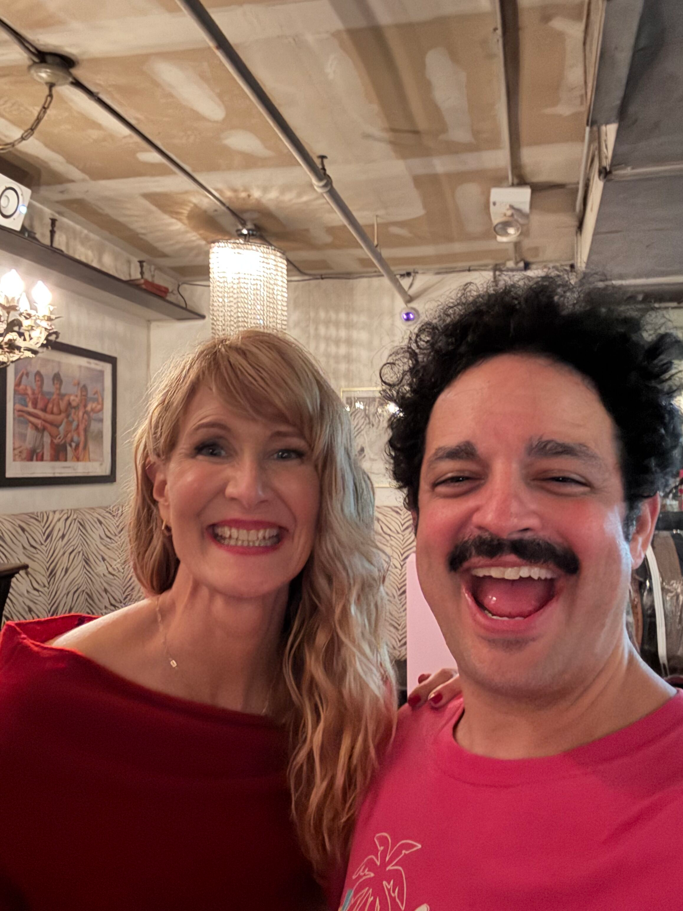A woman with blonde hair and a red top smiling and a man with dark curly hair, mustache, and pink shirt smiling inside a decorated restaurant or cafe with framed artwork and wall lighting.