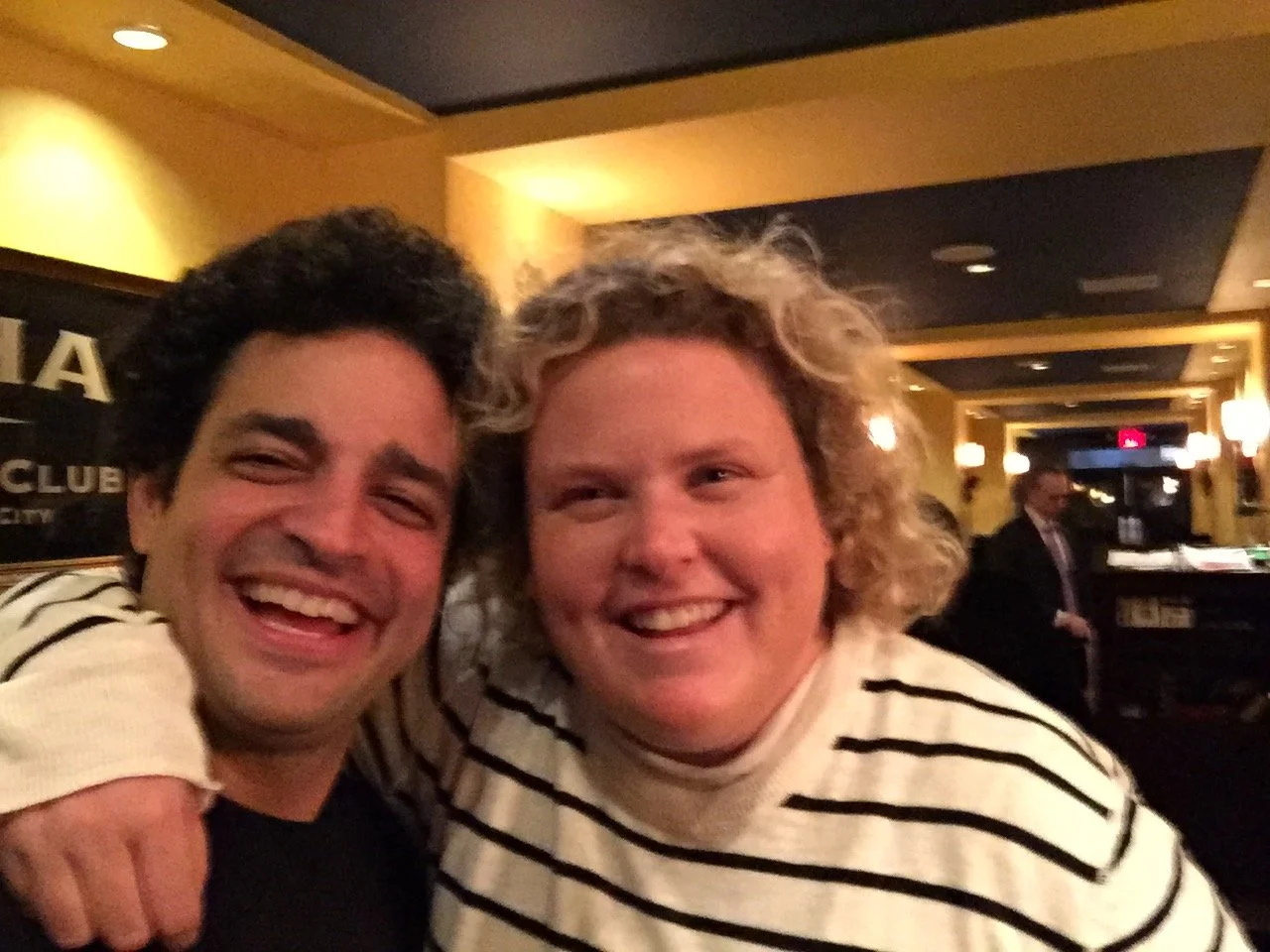 Two people smiling, one with curly black hair wearing a cream sweater with black stripes, the other with blonde curly hair wearing a similar sweater, in a warmly lit indoor setting.