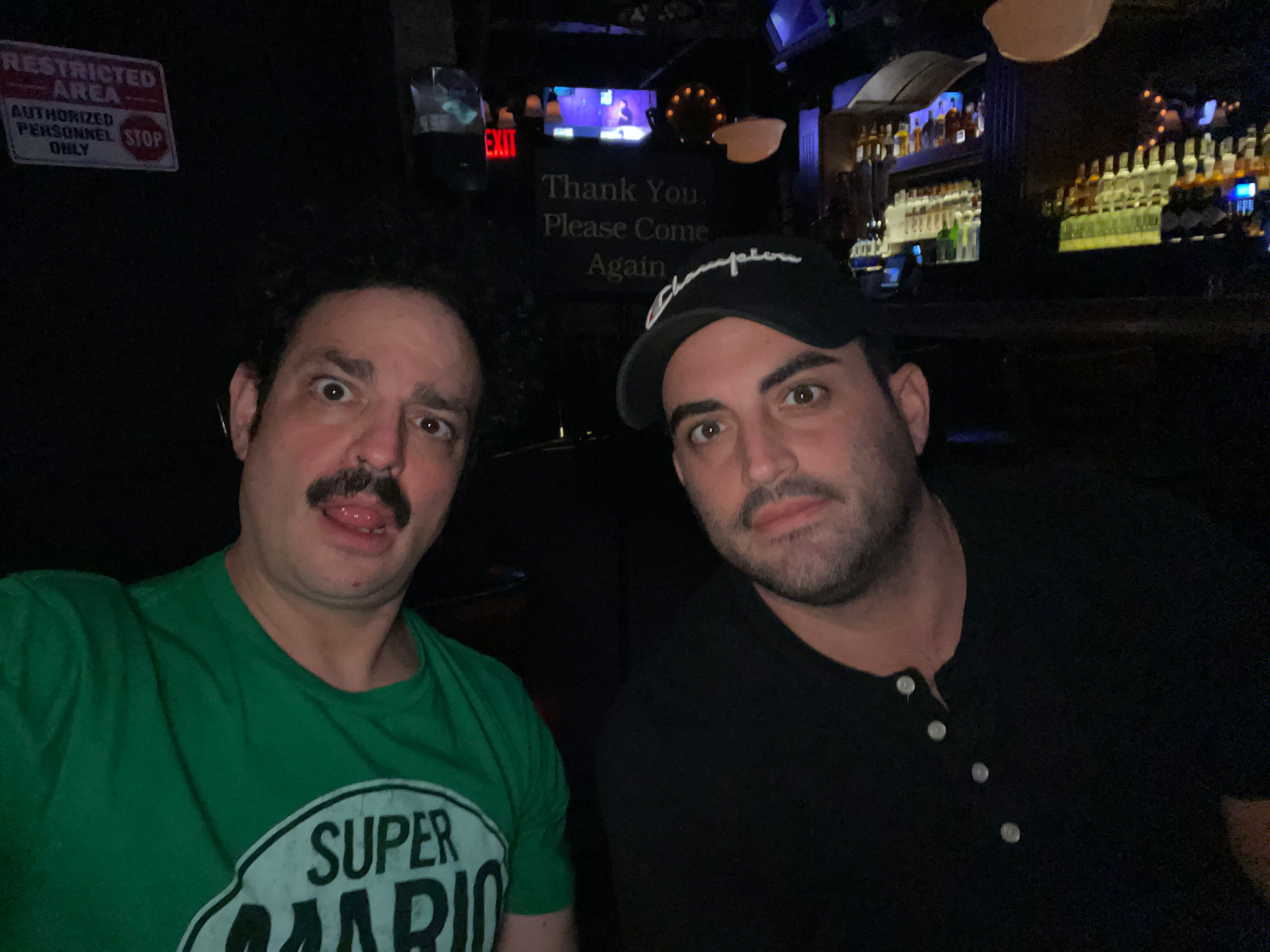 Two men taking a selfie in a dimly lit bar or nightclub. One man is wearing a green shirt with a 'Super' logo, the other is wearing a dark shirt and a baseball cap. The background shows bottles on shelves, descriptions, and television screens. A sign