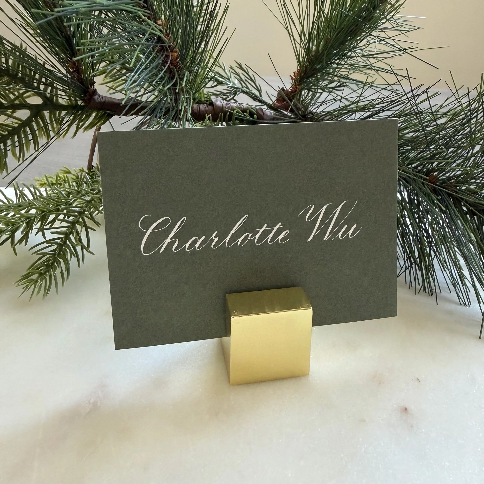 Green holiday place card with a name handwritten in white calligraphy, set against a pine background