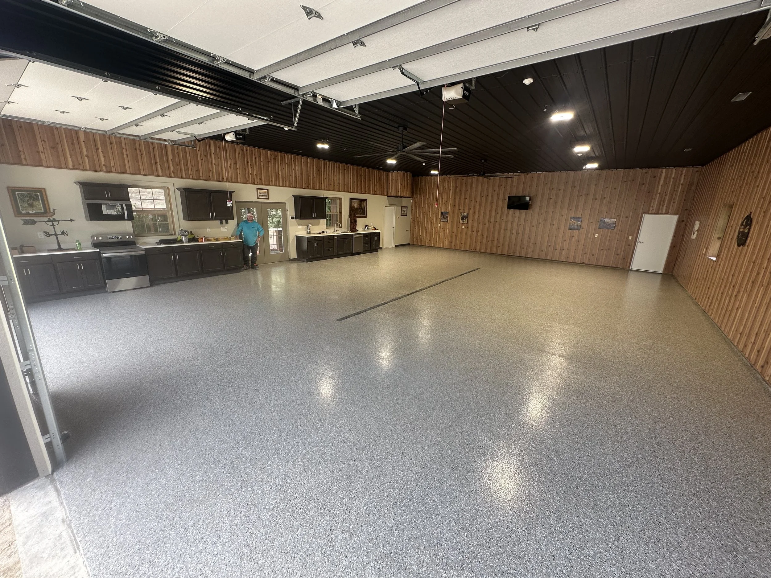 A spacious room with wooden paneling on the walls, a black ceiling, and a floor with a speckled pattern. There is a kitchen area with dark cabinets, a stovetop, and a person standing near the door leading outside. The ceiling features several lights and ceiling fans, and there are mounted speakers and a mounted TV on the walls.