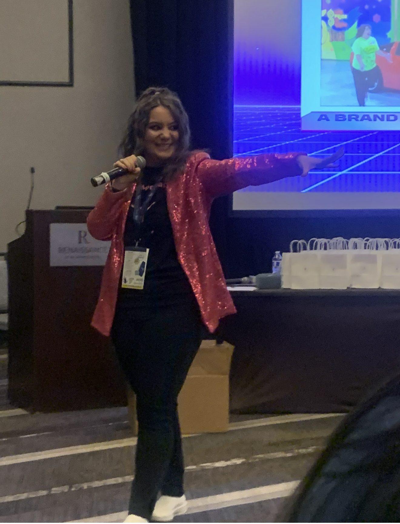 Lisa Moe in a pink sequined jacket holding a microphone and pointing to a game-show slide, hosting her keynote like a game show host