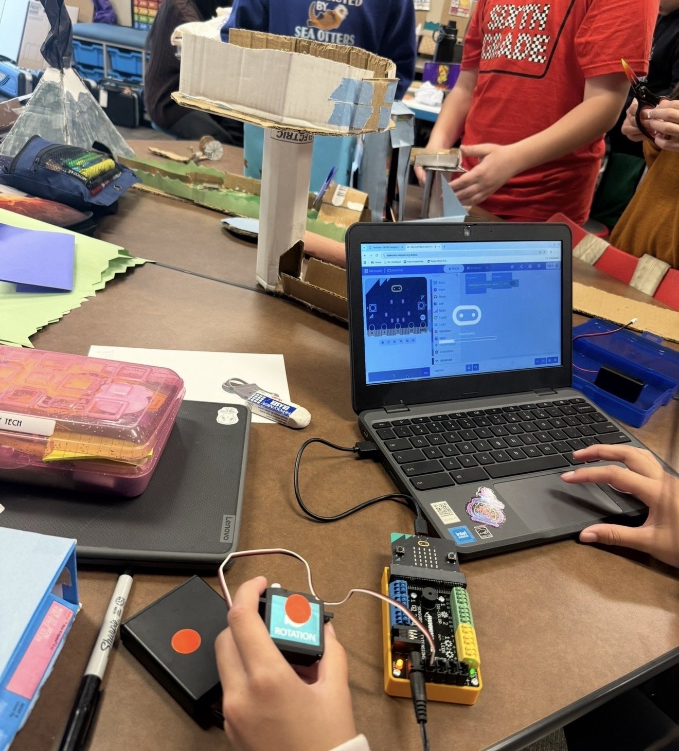 A student coding a micro:bit on a Chromebook, holding a rotation sensor wired to the board, with cardboard engineering projects on the table behind.