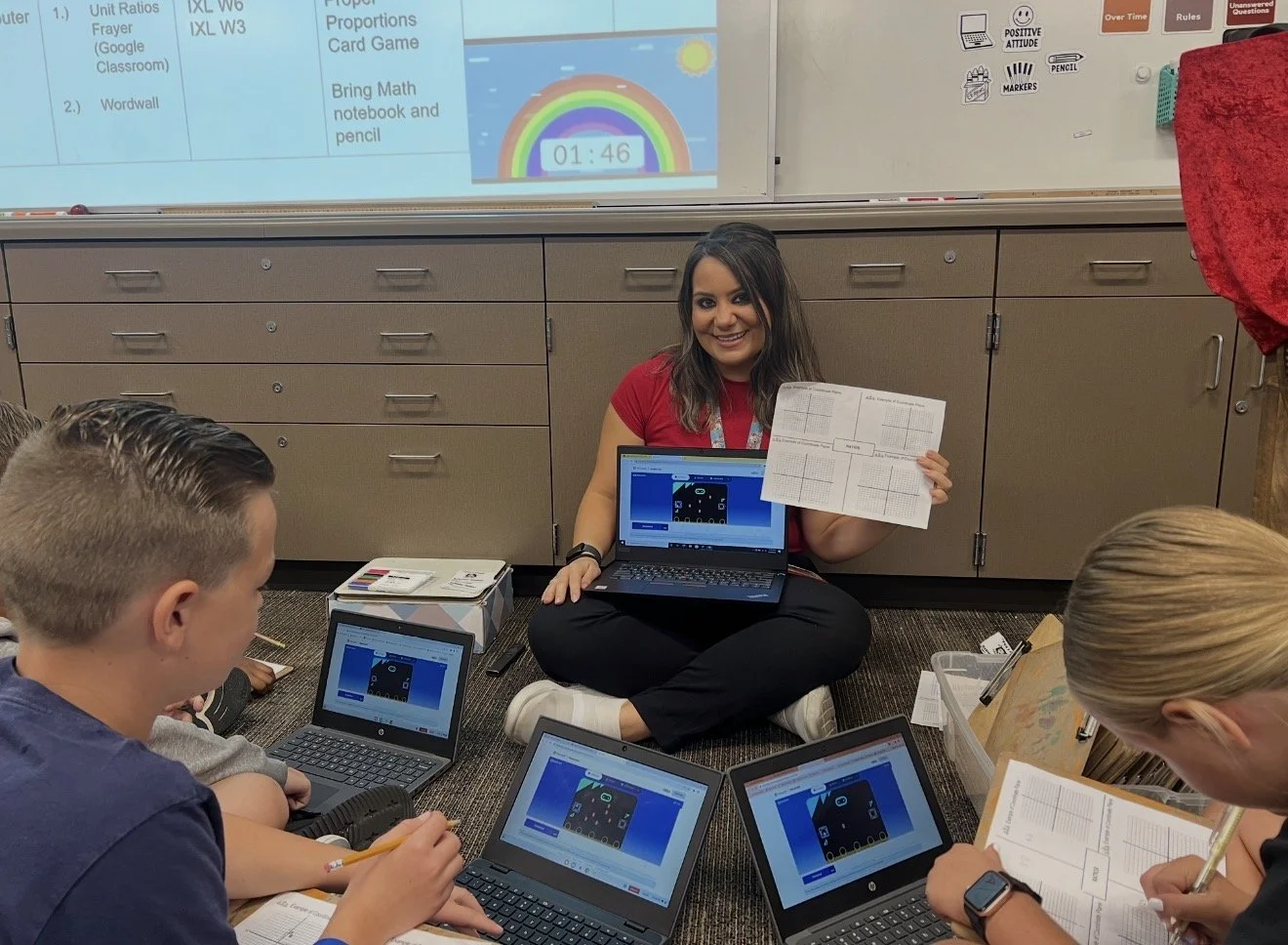 Lisa Moe sitting on the classroom floor with students, holding up a micro:bit coding lesson on her laptop.