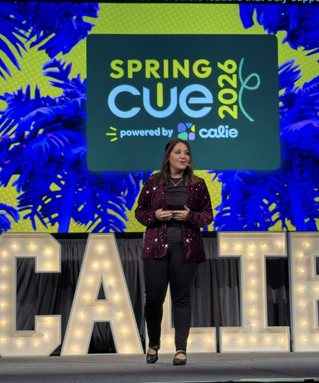 Lisa Moe in a sequined jacket delivering the CALIE Spring CUE 2026 keynote on stage in front of giant marquee letters
