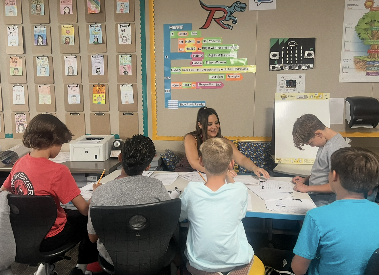 Lisa Moe smiling and working with a small group of students at a classroom table, with a coding-themed leadership board on the wall behind them.
