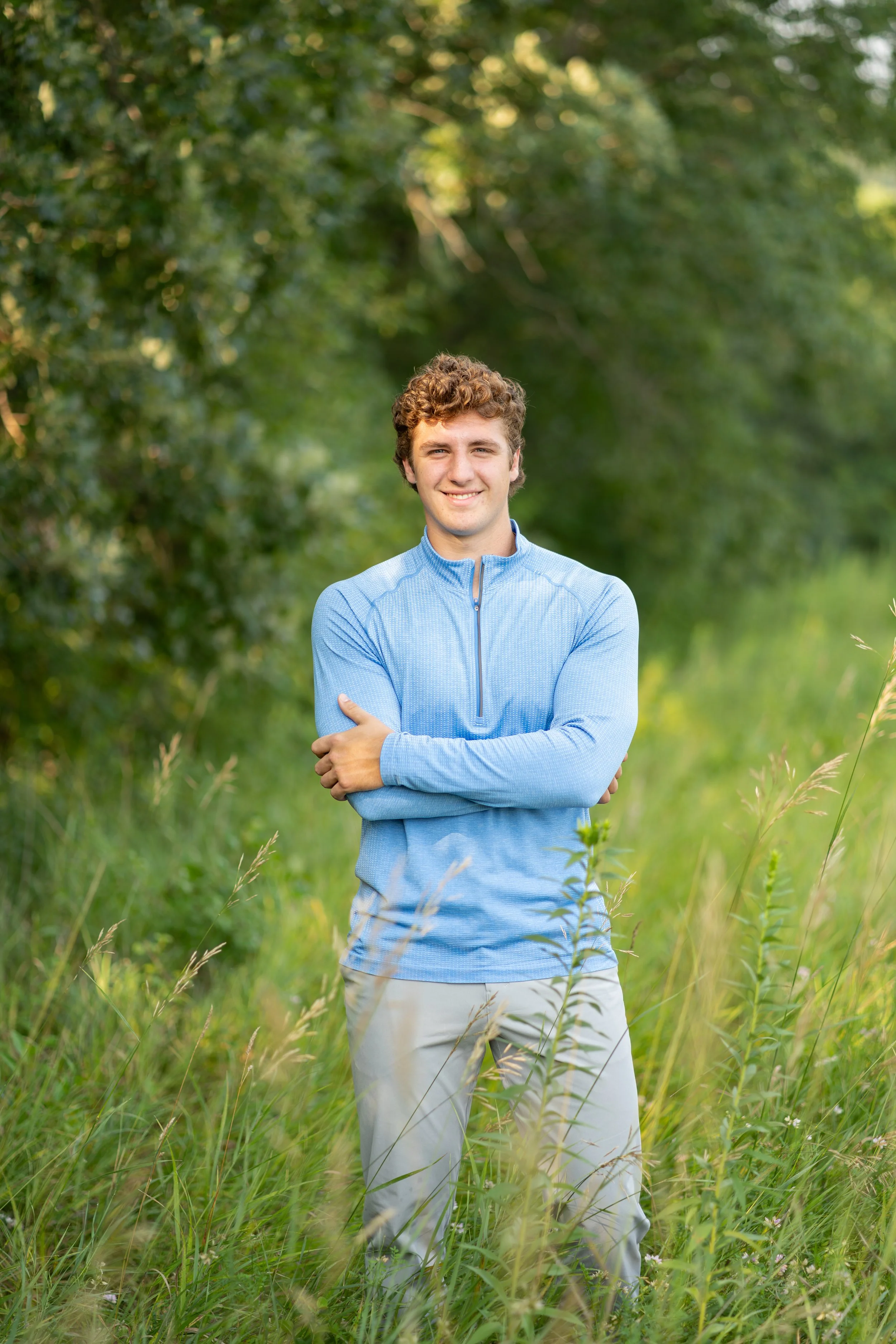 A young man standing in a grassy field with trees in the background, smiling and wearing a blue zip-up jacket and beige pants.