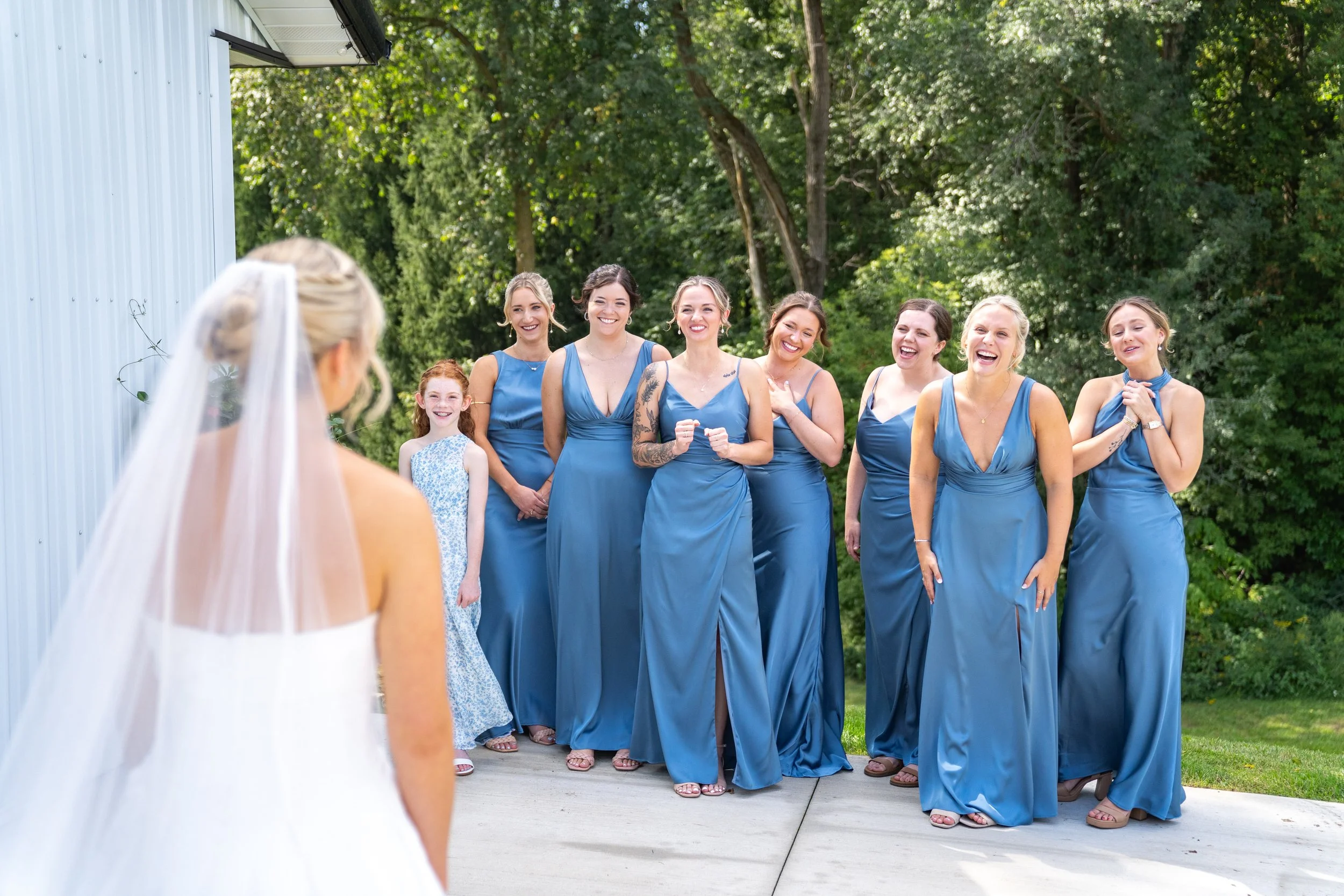 A bride in a white wedding dress with a veil is facing a group of women in blue dresses, smiling outdoors near a garden with trees.