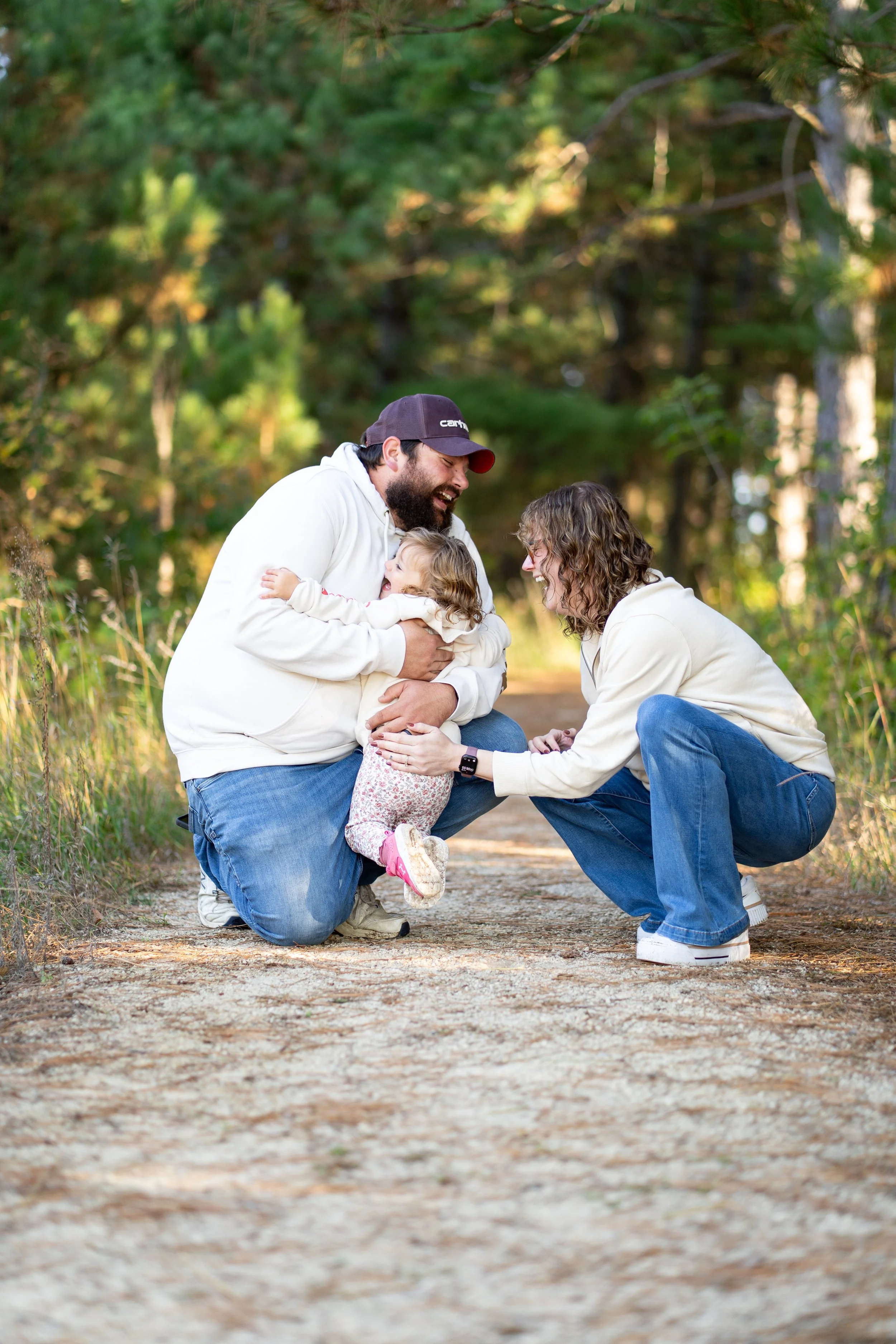 A family of three, two adults and a young girl, enjoying a moment outdoors on a dirt path in a wooded area. The man is kneeling on one knee, holding the girl who is smiling and hugging him, while the woman crouches down, touching the girl's hand and smiling.