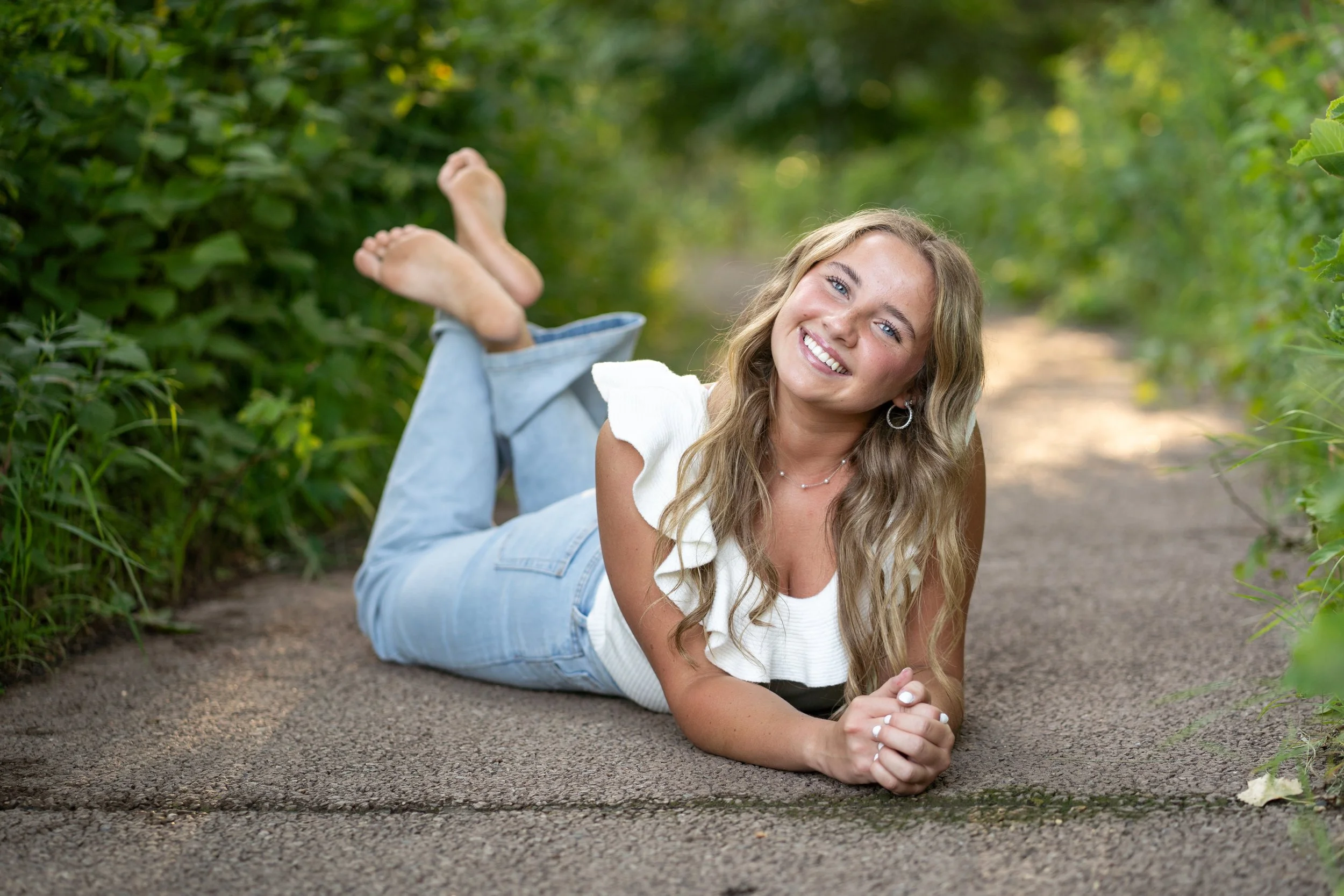 A young woman lying on a dirt path in a forested area, smiling at the camera, with her arms folded under her head and her remaining legs crossed at the ankles, wearing a white top and light blue jeans.