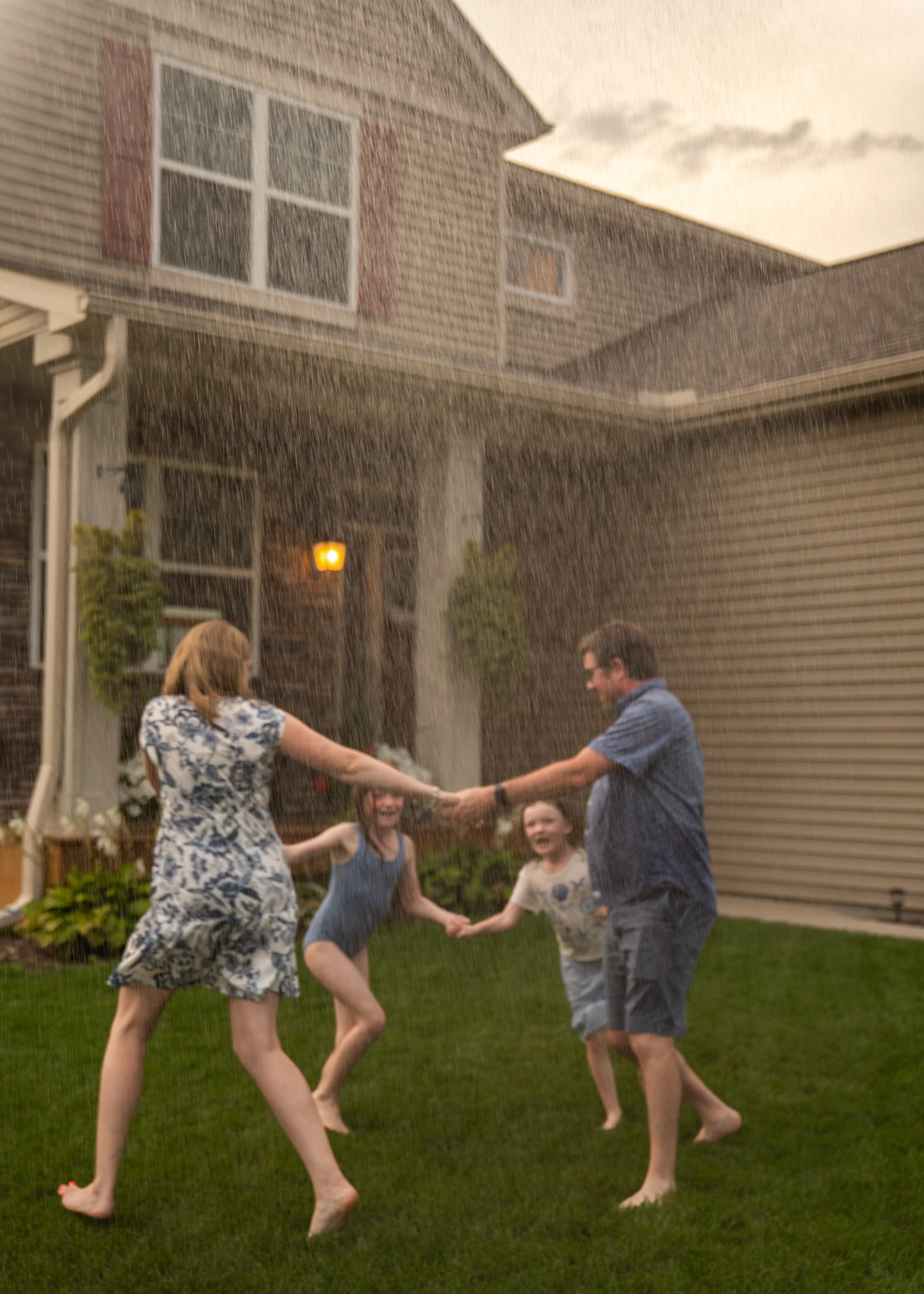 People holding hands and dancing in a backyard during a rainstorm, with a house, a fence, and a cloudy sky in the background.