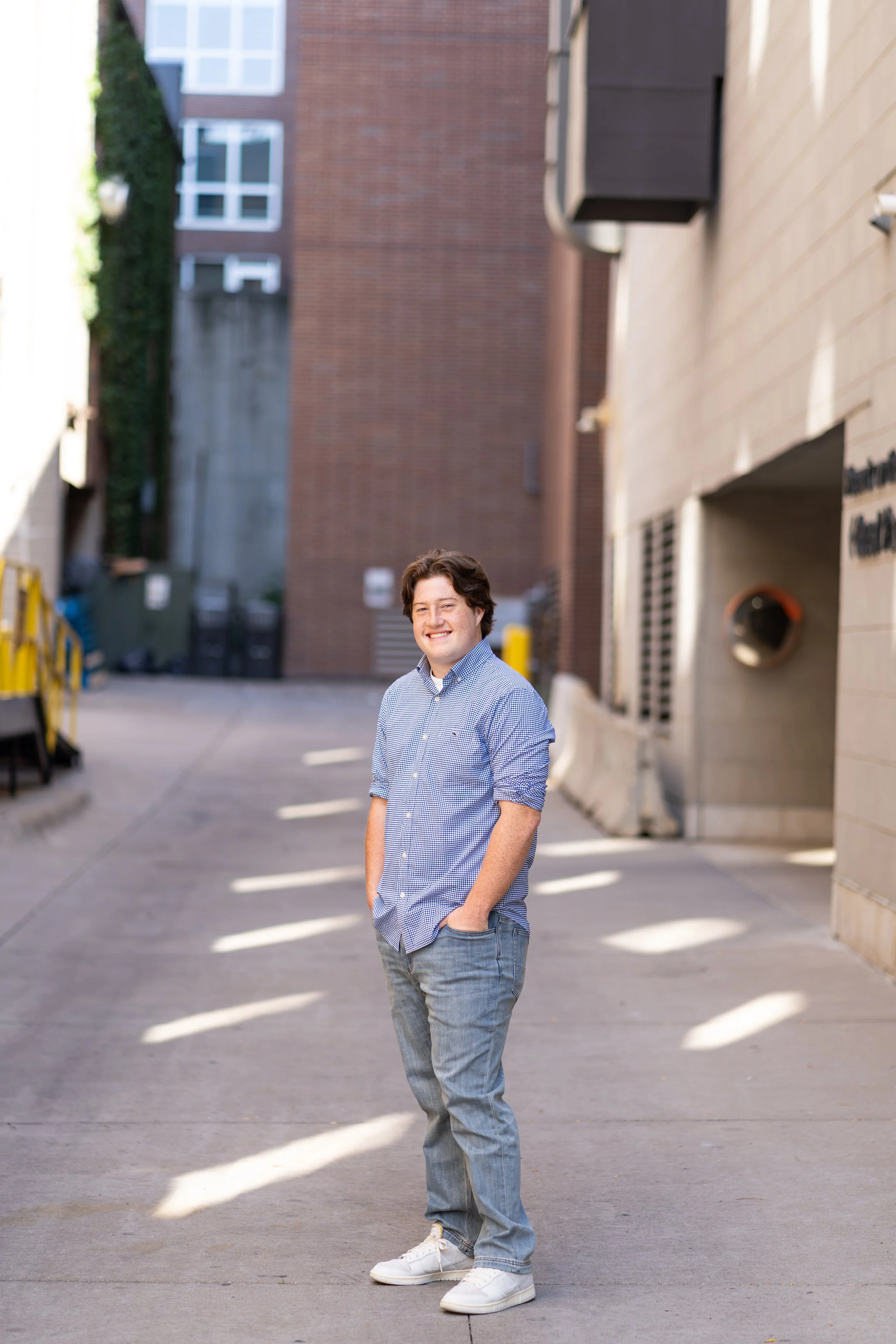 A young man with brown hair, wearing a blue checkered shirt, light blue jeans, and white sneakers, stands outdoors on a paved pathway, smiling with hands in pockets, with brick and concrete buildings in the background.