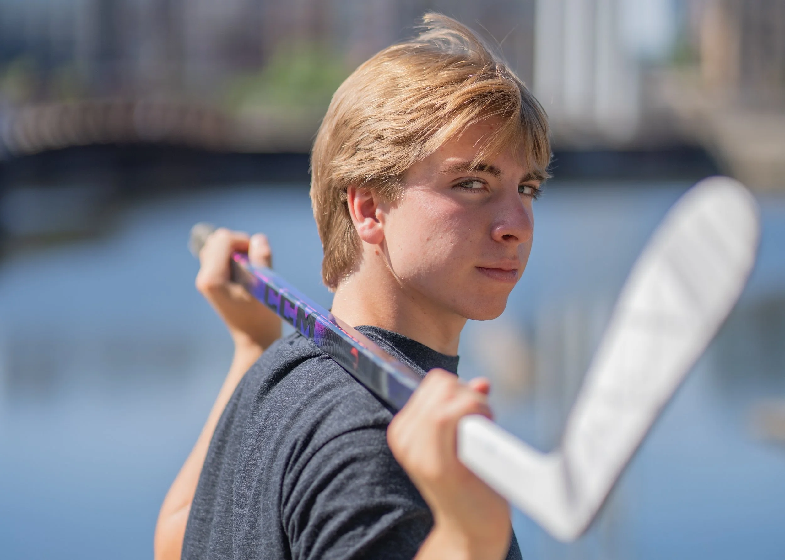 A young boy with light brown hair and a gray t-shirt holding a hockey stick outdoors near a body of water.