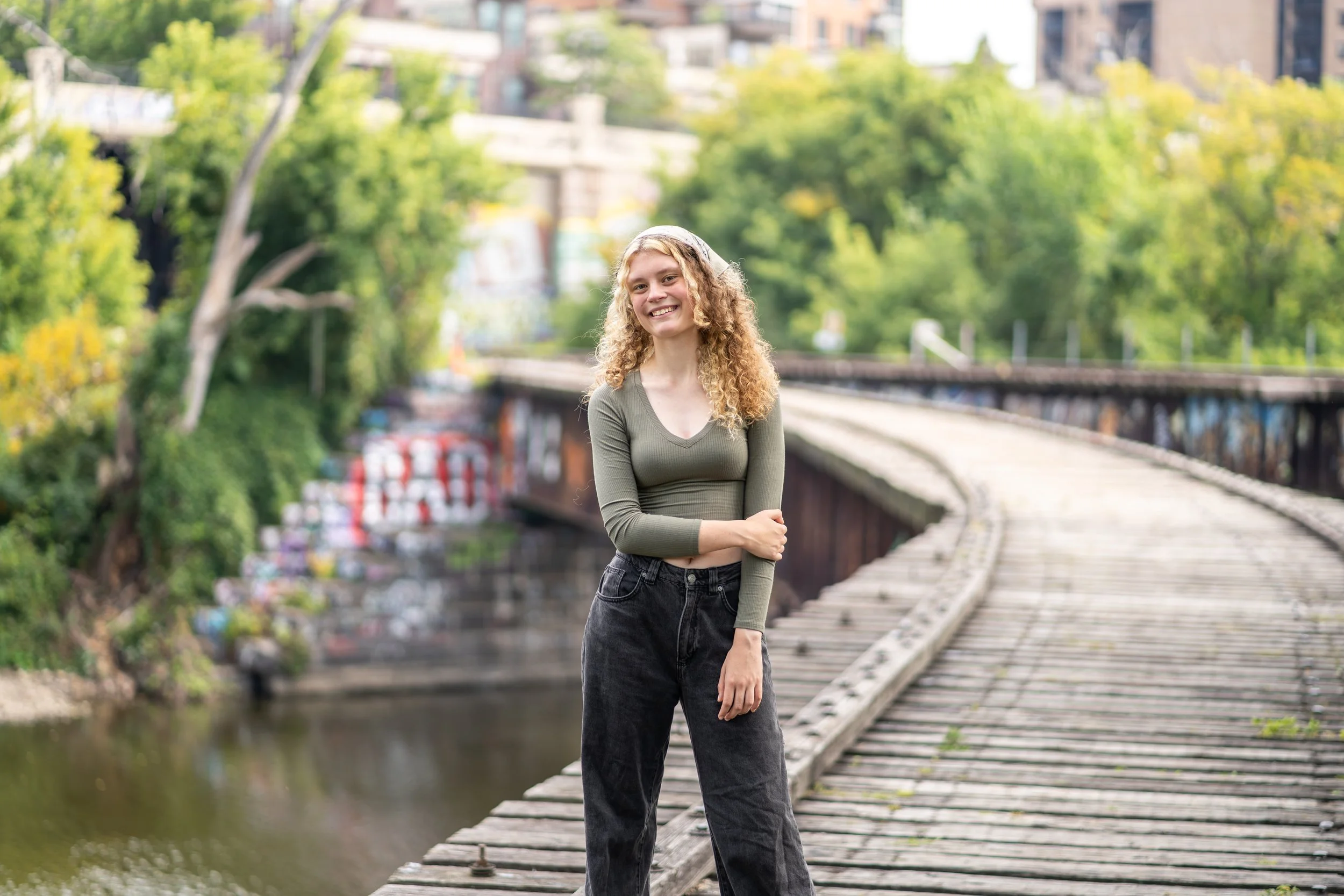 A young woman with curly hair smiling, standing on a wooden bridge over a river, with green trees and blurred buildings in the background.