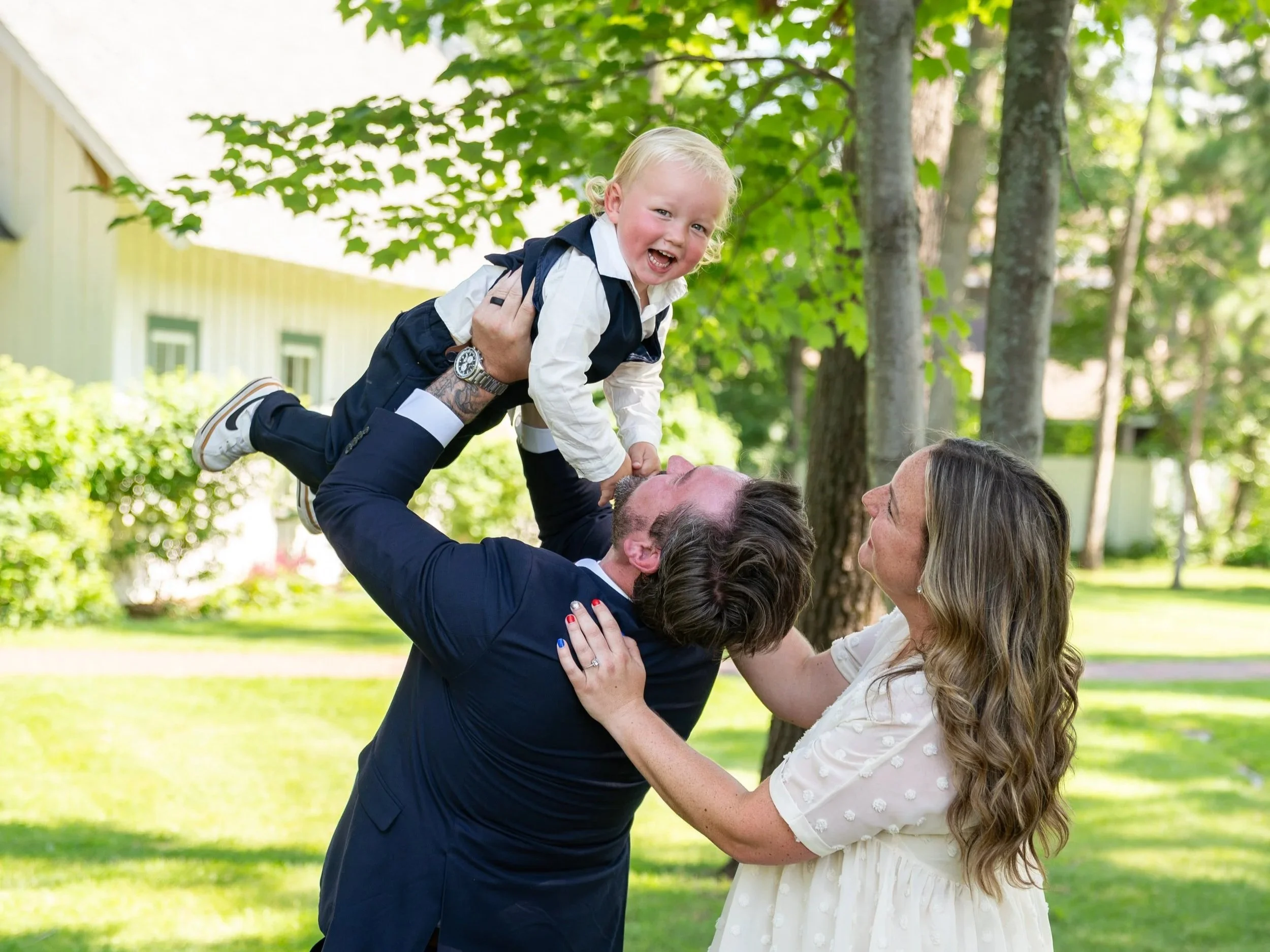 A man lifting a young girl in an outdoor park, both smiling and enjoying a sunny day with green trees in the background.