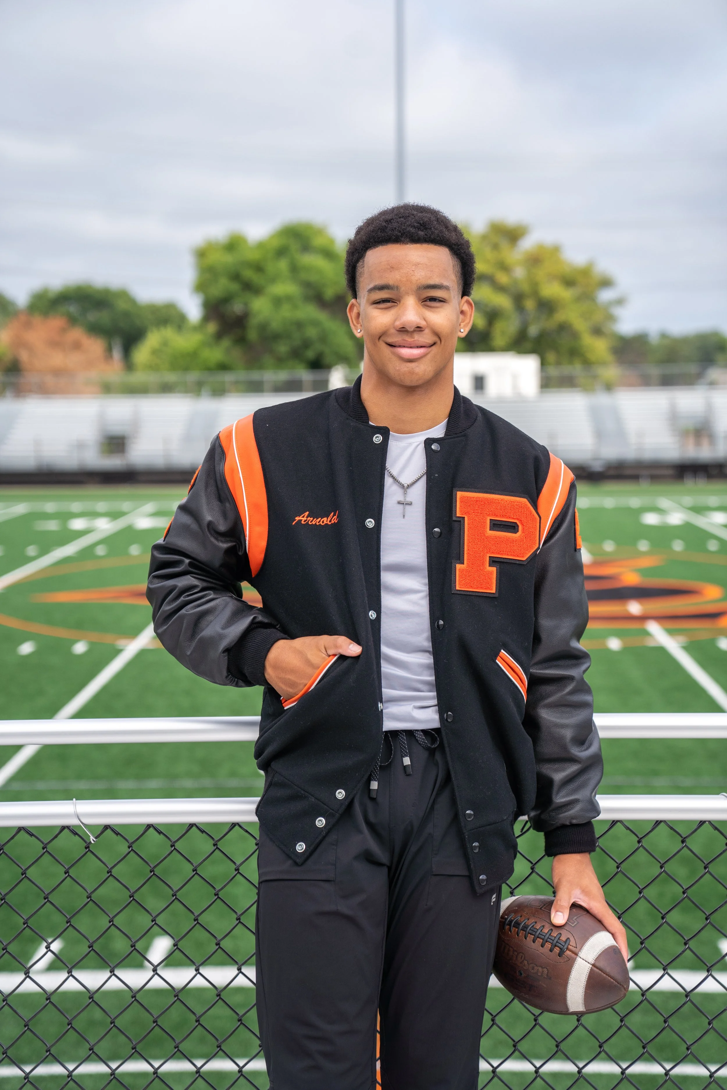 Young man standing on a football field with a football in his right hand, wearing a black varsity jacket with orange accents and a letter P, and standing in front of a chain-link fence with trees and bleachers in the background.