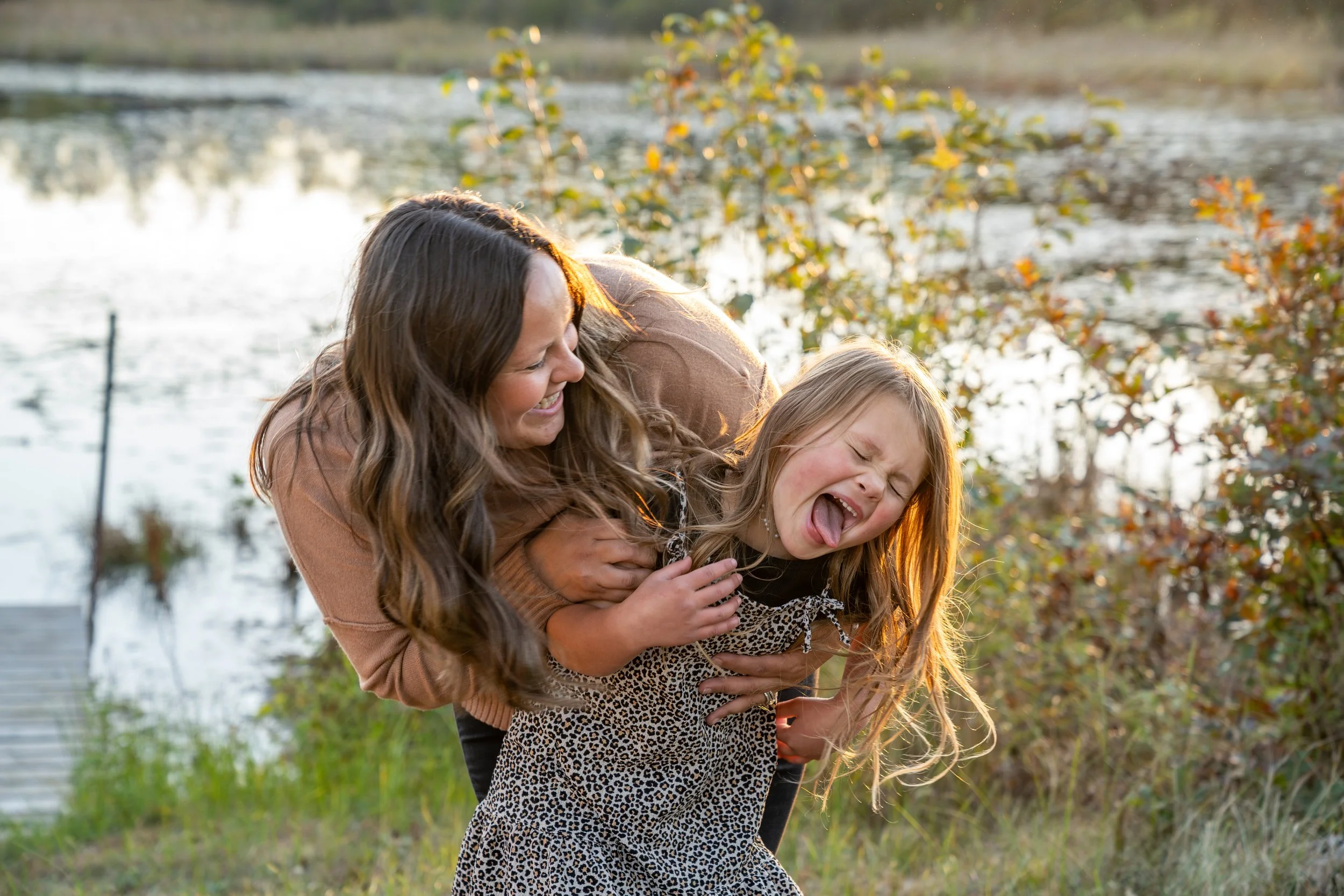 A woman and a young girl playfully laughing outdoors near a body of water, surrounded by trees with autumn-colored leaves.