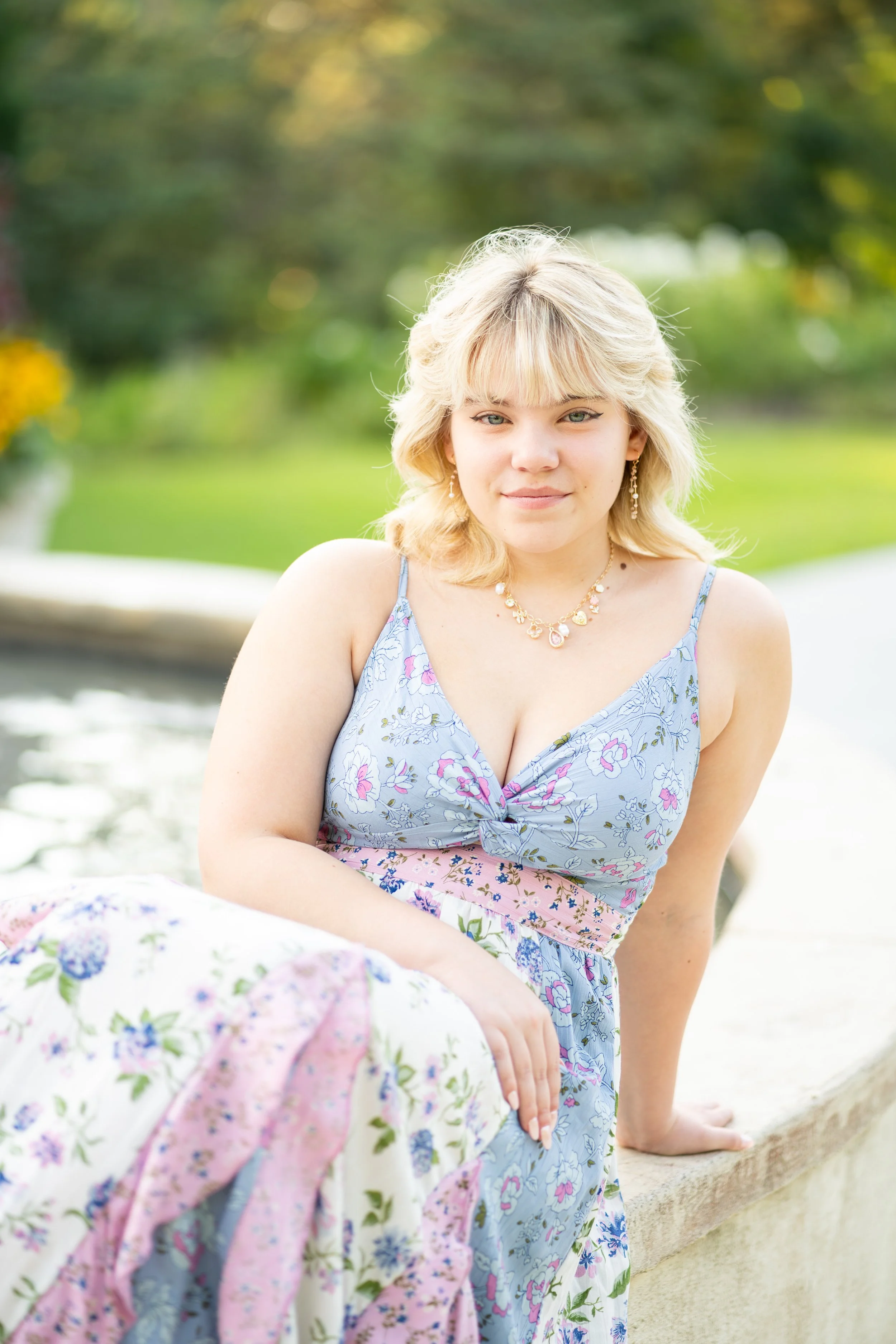 A young woman with blonde hair sitting outdoors near a stone ledge with green foliage background, wearing a floral dress and jewelry.
